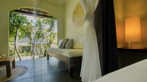 Una habitación con cama, sofá, una mesita y una lámpara. Se ve un paisaje con árboles y un edificio por la ventana.