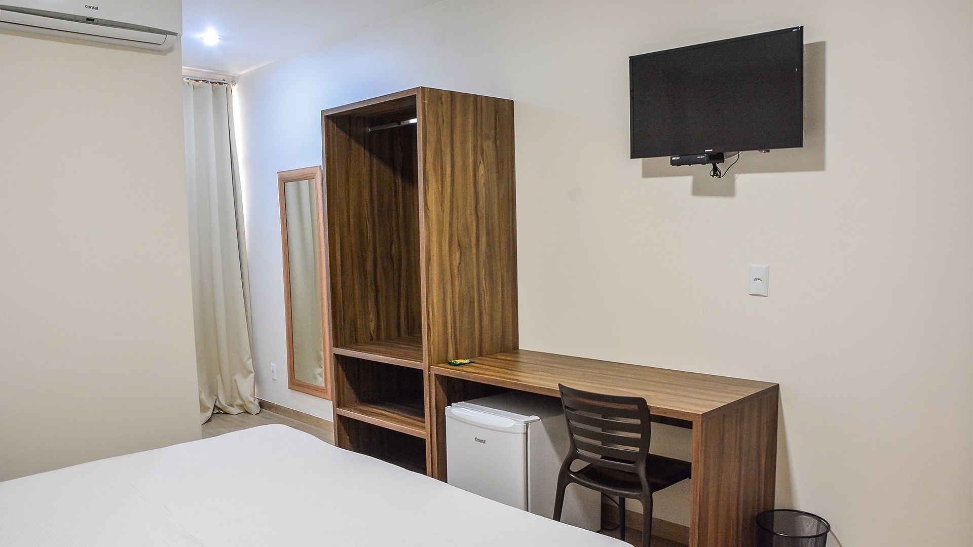 A bedroom with a bed, a desk, a chair, a small refrigerator, and a TV mounted on the wall.