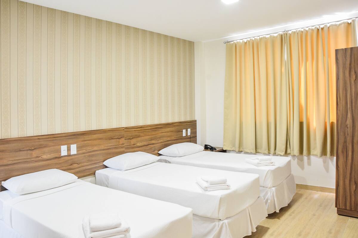 A hotel room with three single beds, wooden headboards, and a large window with curtains.