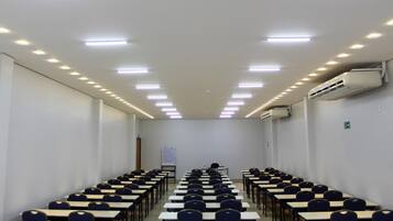 A conference room with rows of tables and chairs, ceiling lights, and an air conditioning unit.