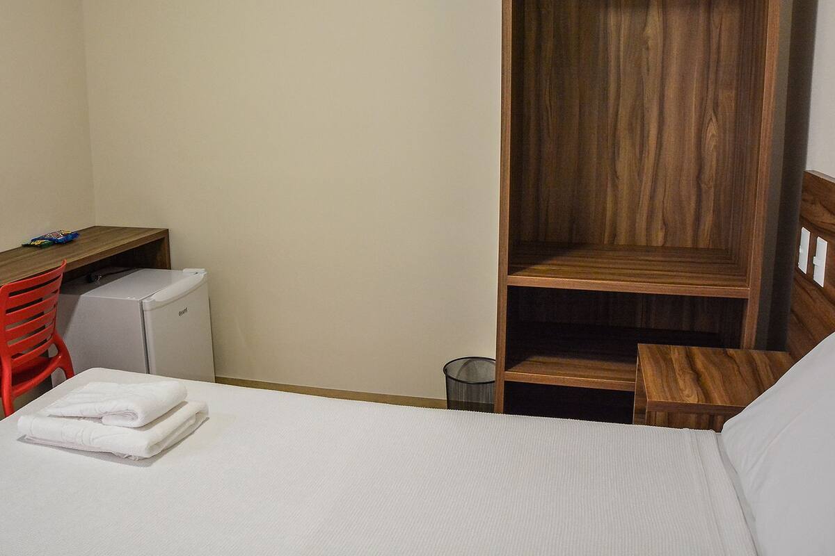 A small, compact room with a bed, a desk, a chair, a small refrigerator, and a wooden wardrobe.