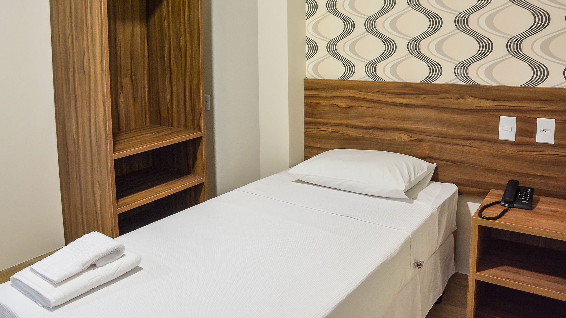 A single bed with a white mattress and pillow, a wooden headboard, a bedside telephone, and a wooden wardrobe with two shelves.
