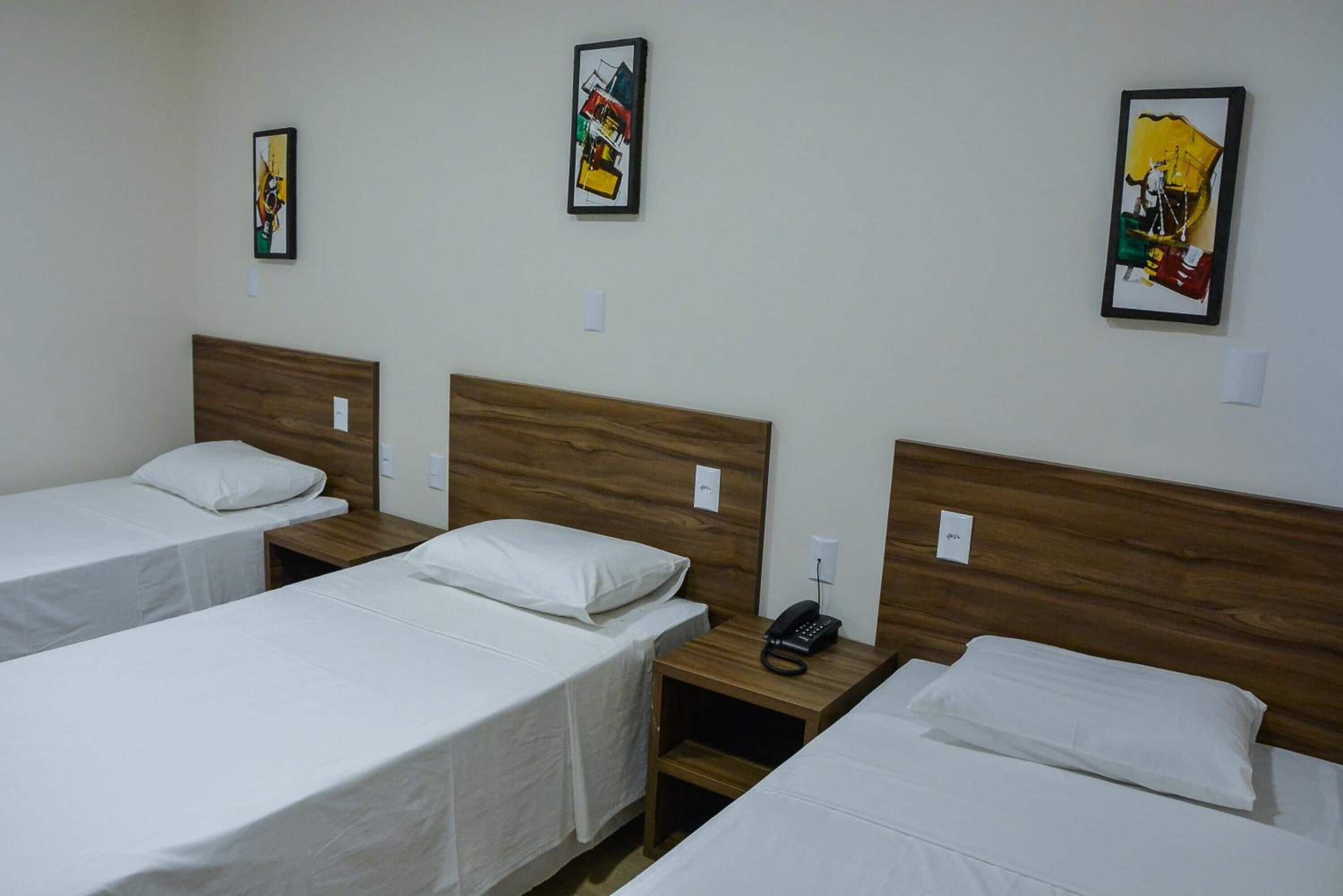 Une chambre d’hôtel avec trois lits simples, des têtes de lit en bois, du linge de lit blanc et une table de chevet avec un téléphone.