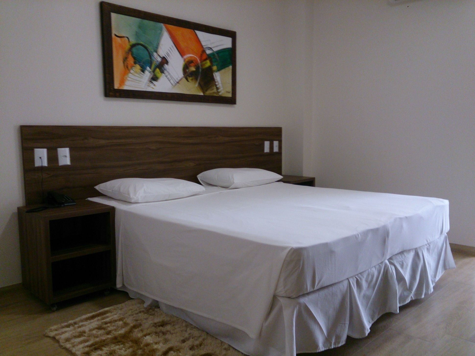 Cama bem arrumada com roupa de cama branca, cabeceira de madeira e uma pequena mesa de cabeceira com abajur.