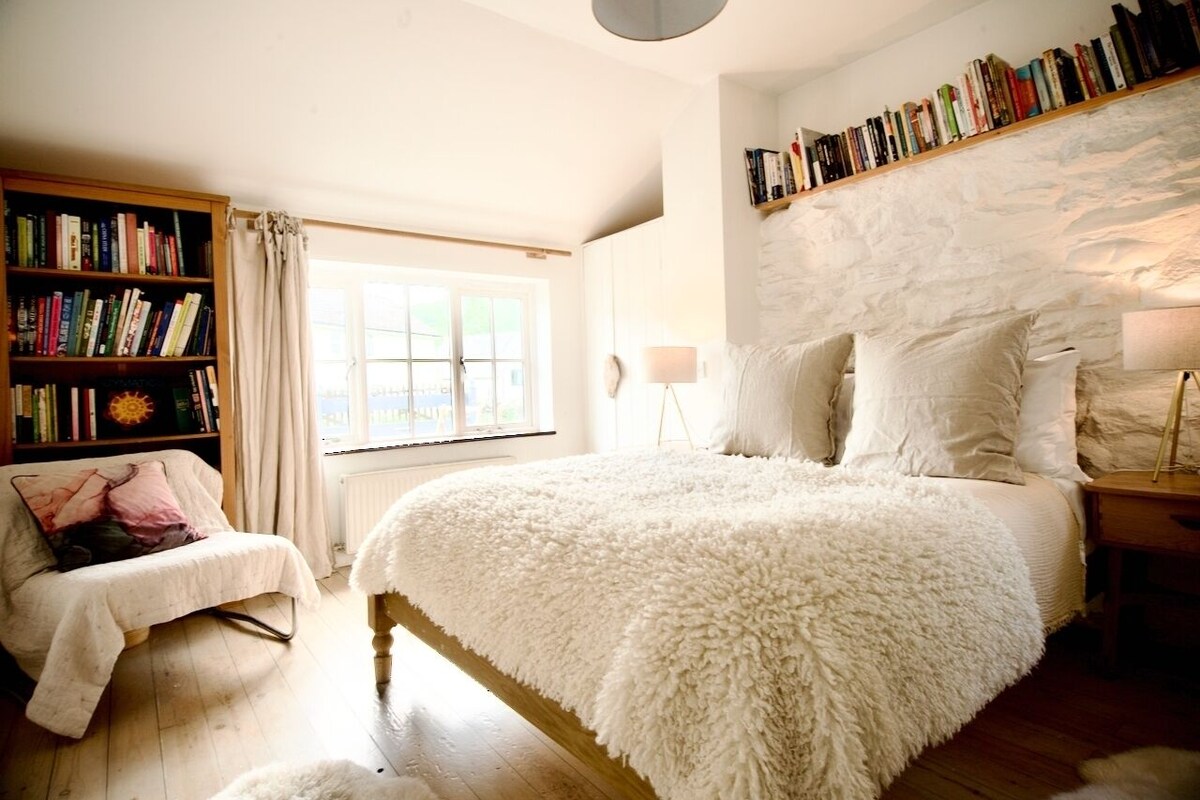 A bedroom with a large bed, a bookshelf, a window, and a white headboard.