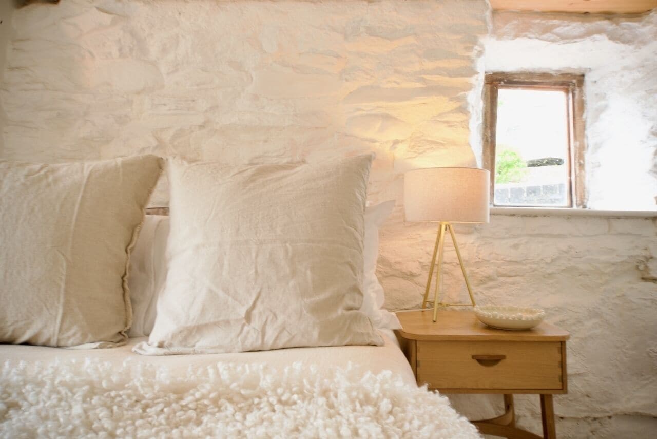 Un dormitorio con pared de piedra, cama con ropa de cama blanca, mesita de noche con lámpara y ventana con vistas.