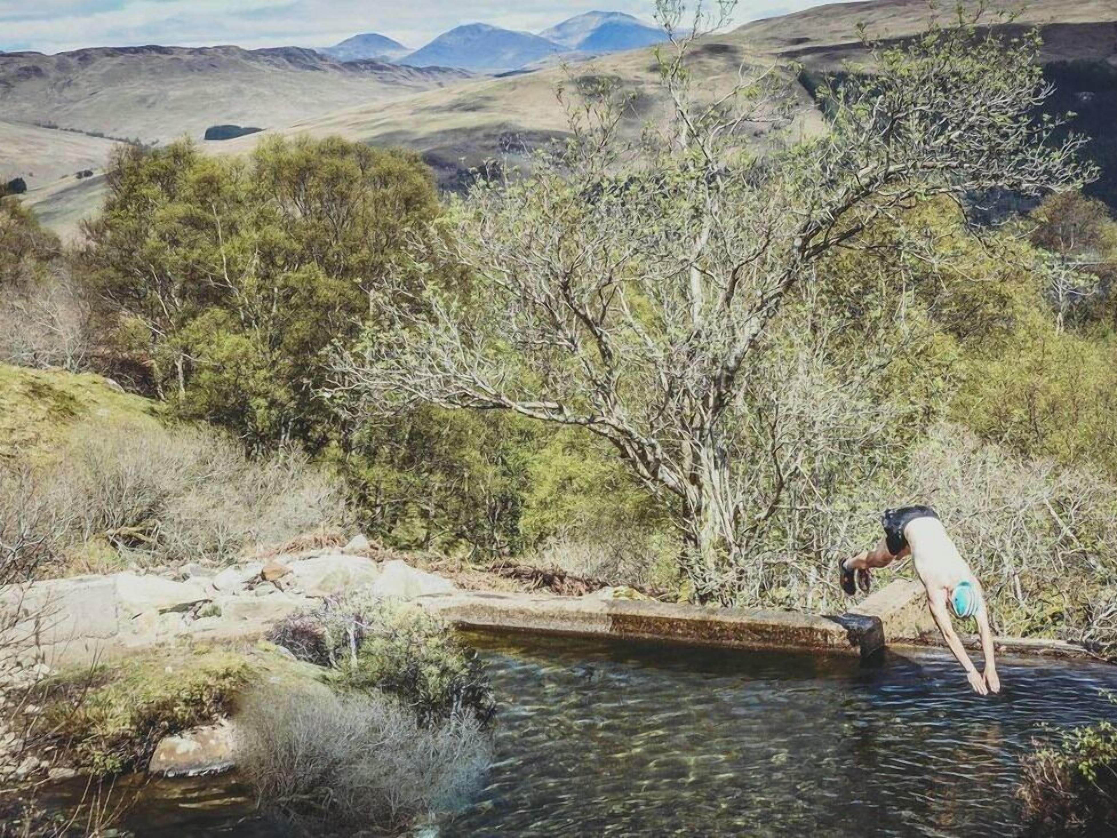 Una persona cerca de una masa de agua en un paisaje natural con colinas al fondo.
