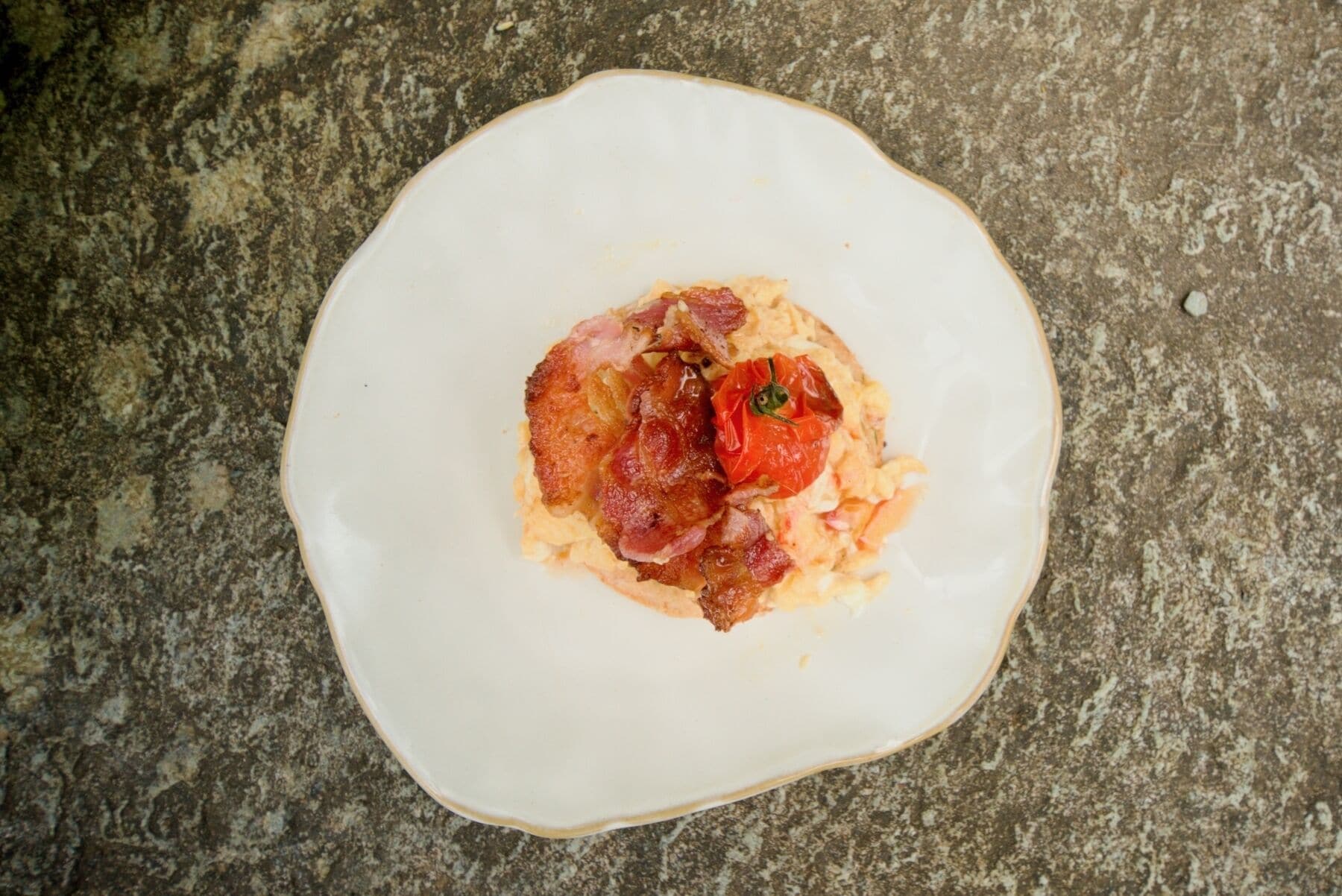 Ein weißer Teller mit Rührei, Speck und einer Tomate auf einer Granitabdeckung.