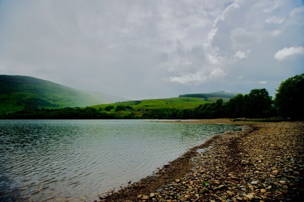 Un lac bordé d’une plage de galets, avec des collines verdoyantes en arrière-plan.