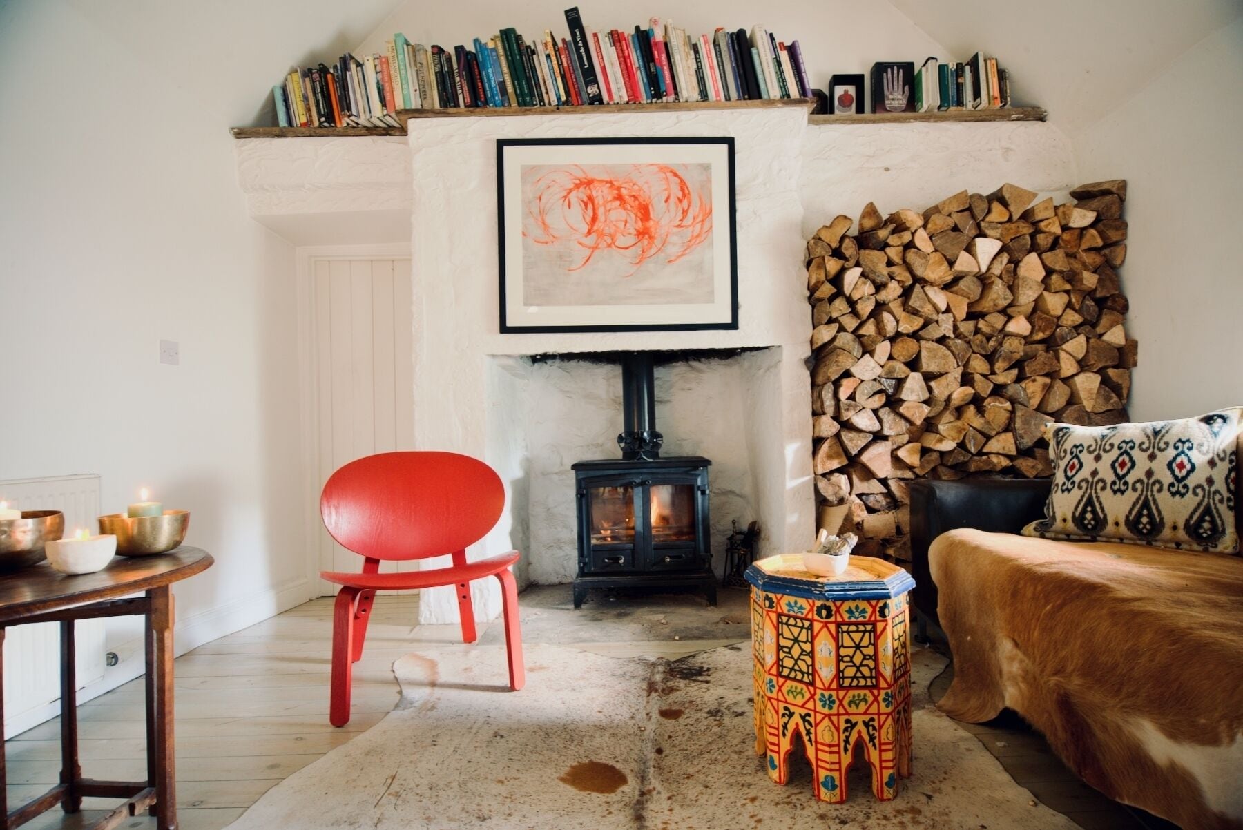 Una sala de estar acogedora con un sillón rojo, un taburete pequeño, una chimenea, una estantería y un cuadro abstracto enmarcado.
