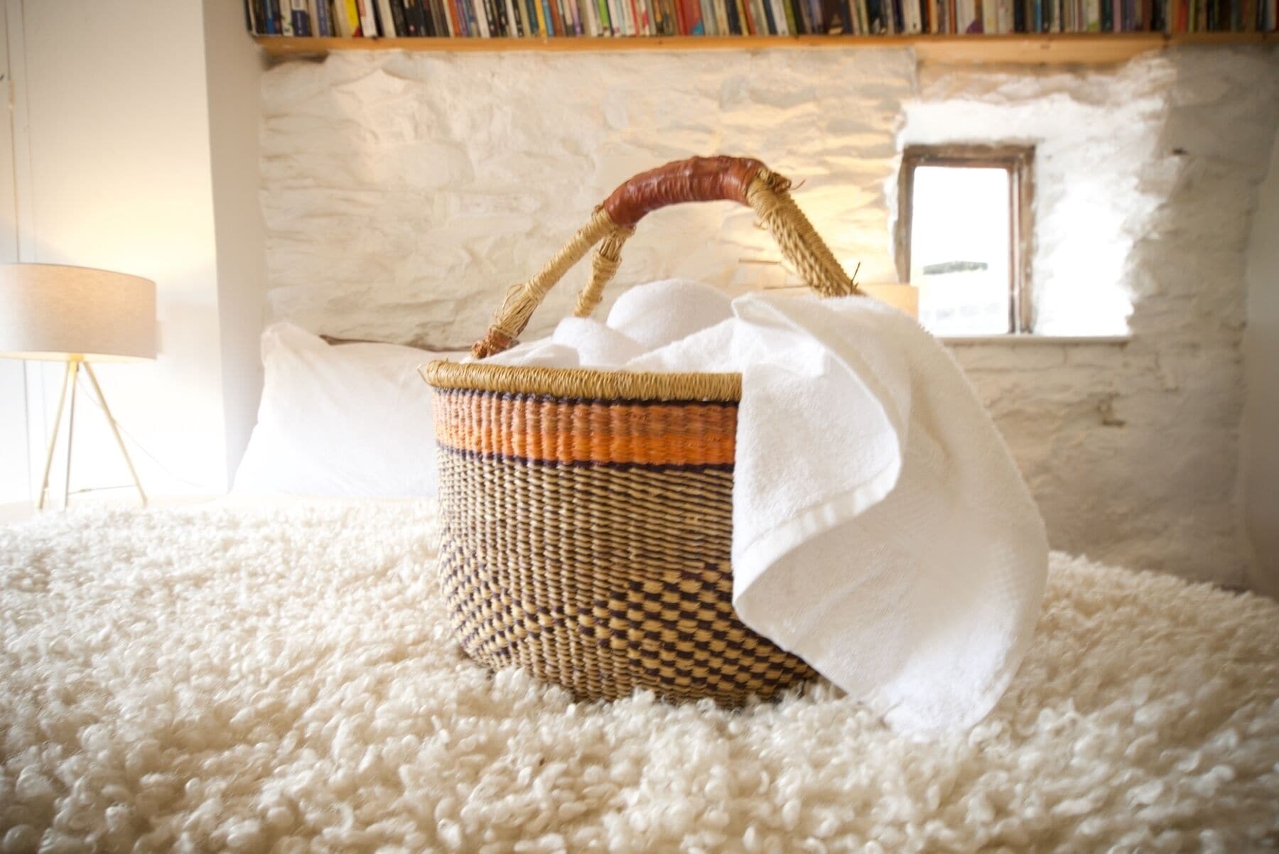 A wicker basket with a handle on a white carpet, a lamp, and a bookshelf in the background.