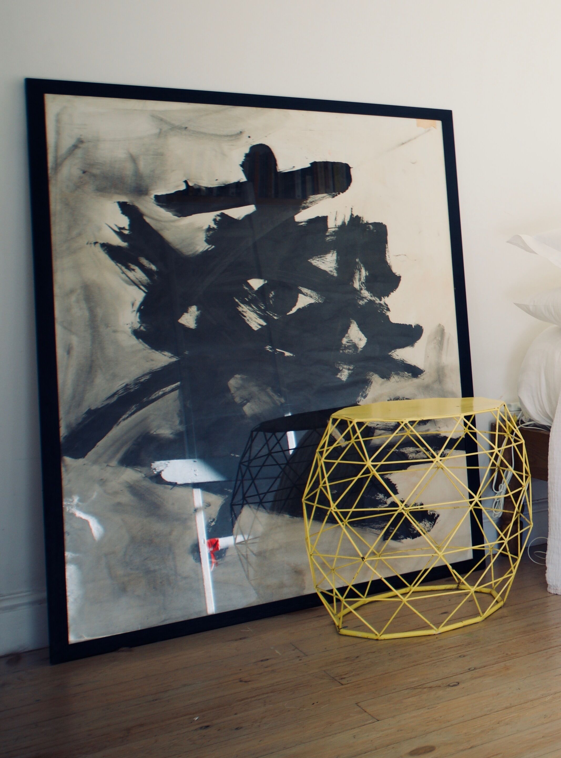 A large abstract painting with a black frame on a wall, a geometric yellow chair, and a wooden floor.