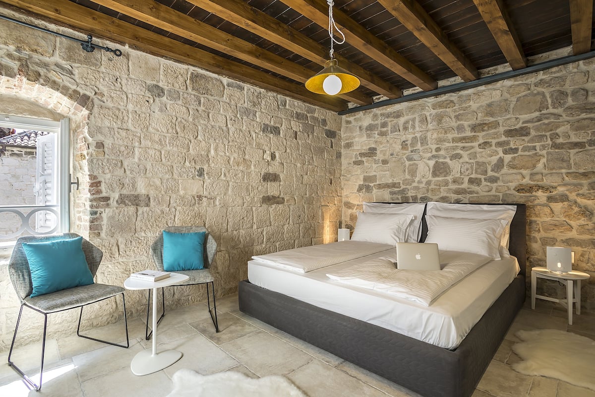 A bedroom with a stone wall, a bed with white linens, a laptop on the bed, and a small table with a chair.