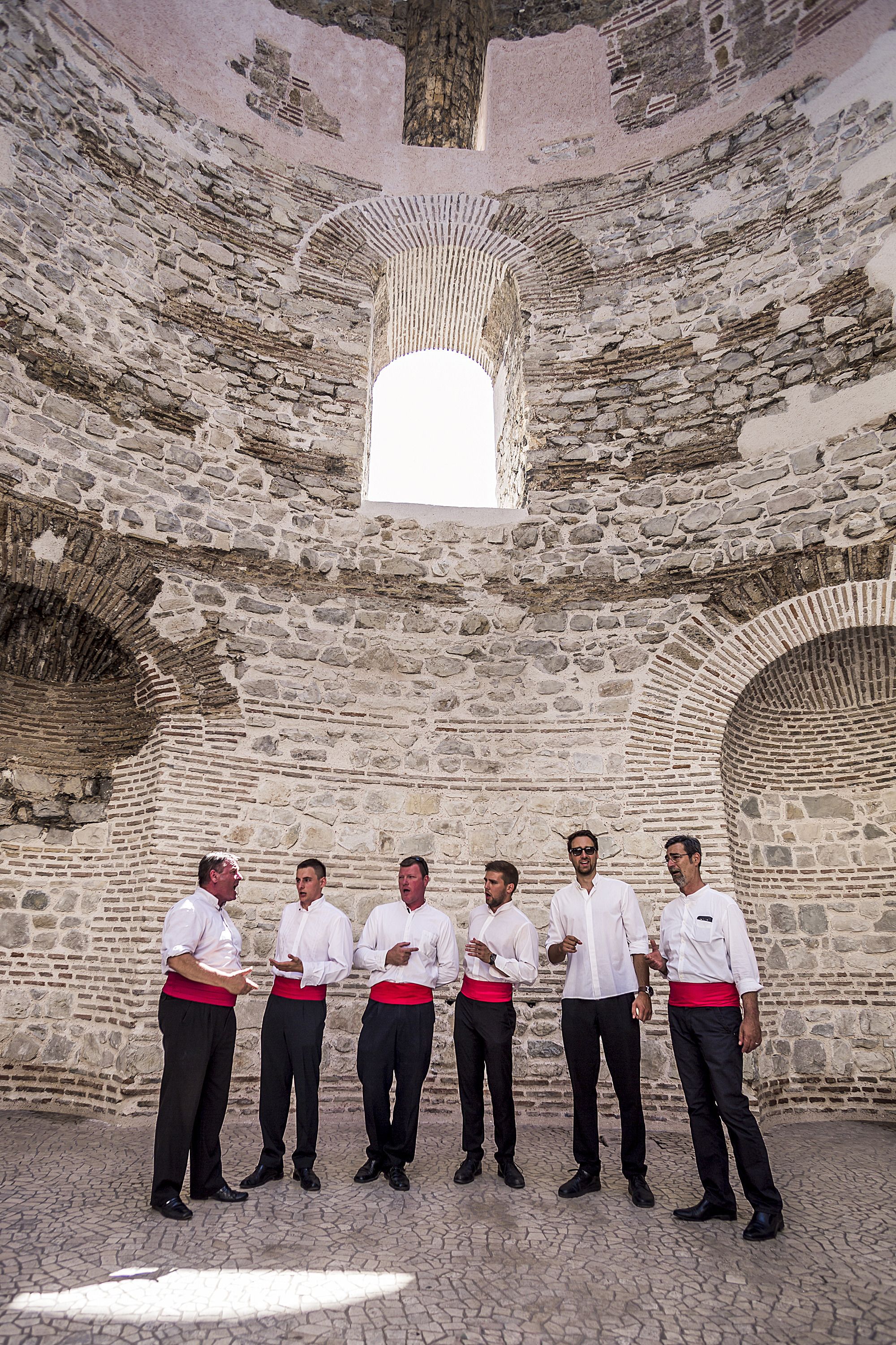 Un groupe d’hommes vêtus de chemises blanches et de pantalons noirs se tient dans une cour bordée de murs en pierre et dotée d’ouvertures en arc de cercle.