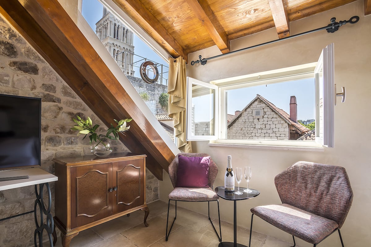 A cozy room with a stone wall, wooden beams, a television, a cabinet, a chair, a small table with a bottle and glasses, and a window with a view of a building.