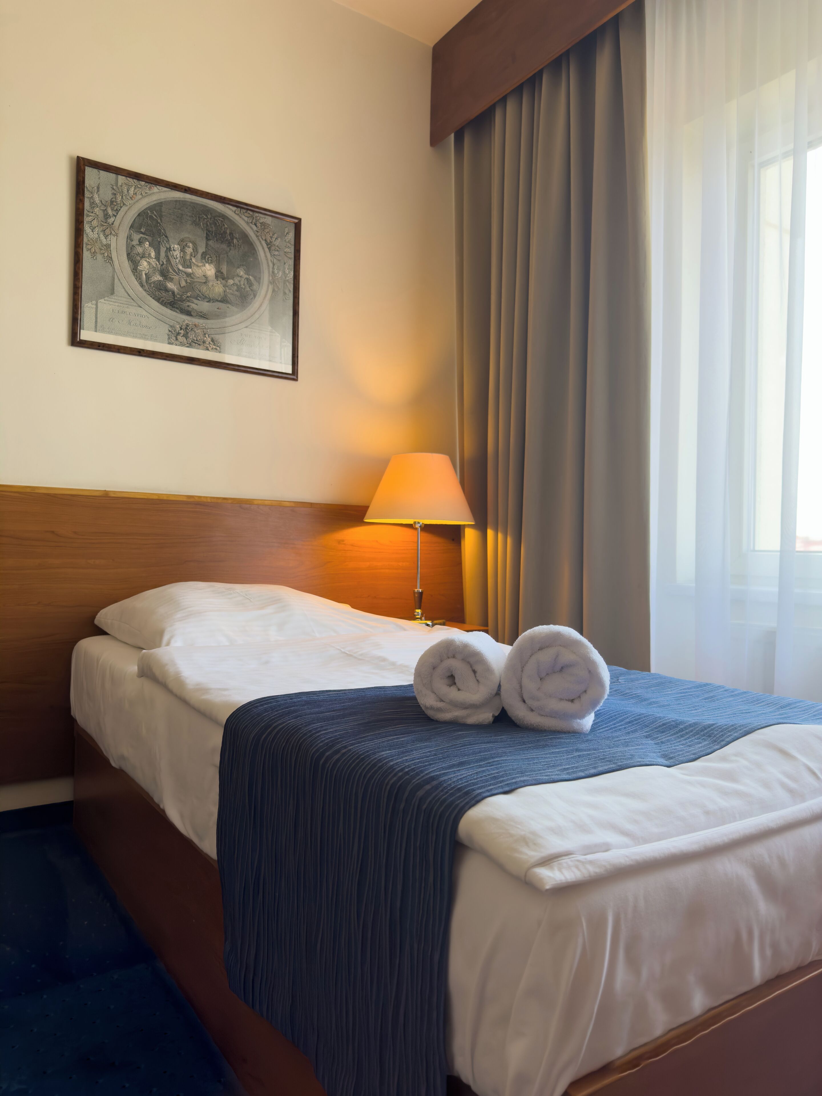 Een hotelkamer met een net opgemaakt bed, een lamp, een ingelijst schilderij aan de muur en een raam met gordijnen.