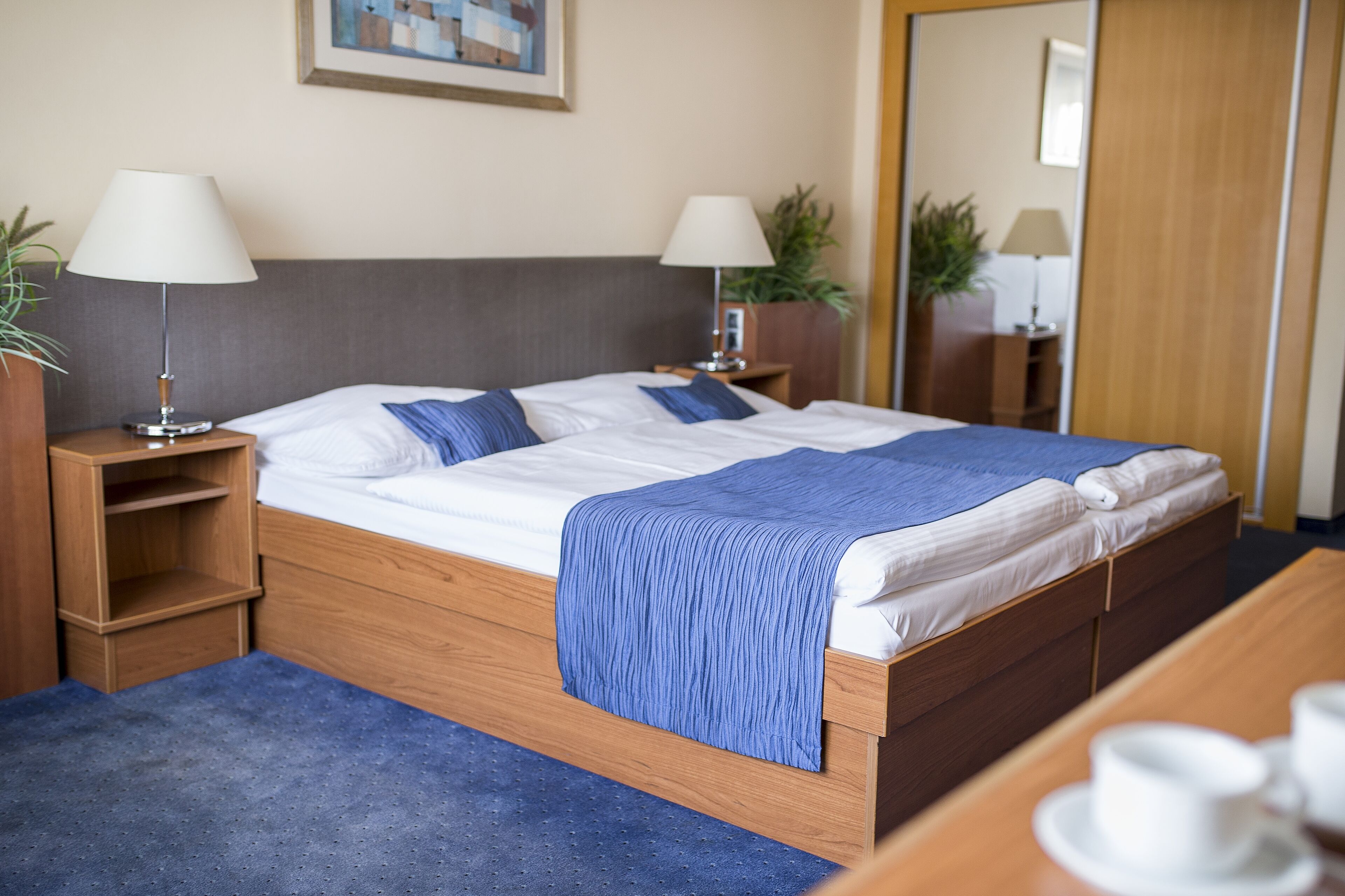 Hotellihuone, jossa on puinen sänky, kaksi yöpöytää valaisimineen, sininen peitto ja pieni puinen sivupöytä, jonka päällä on kasvi.