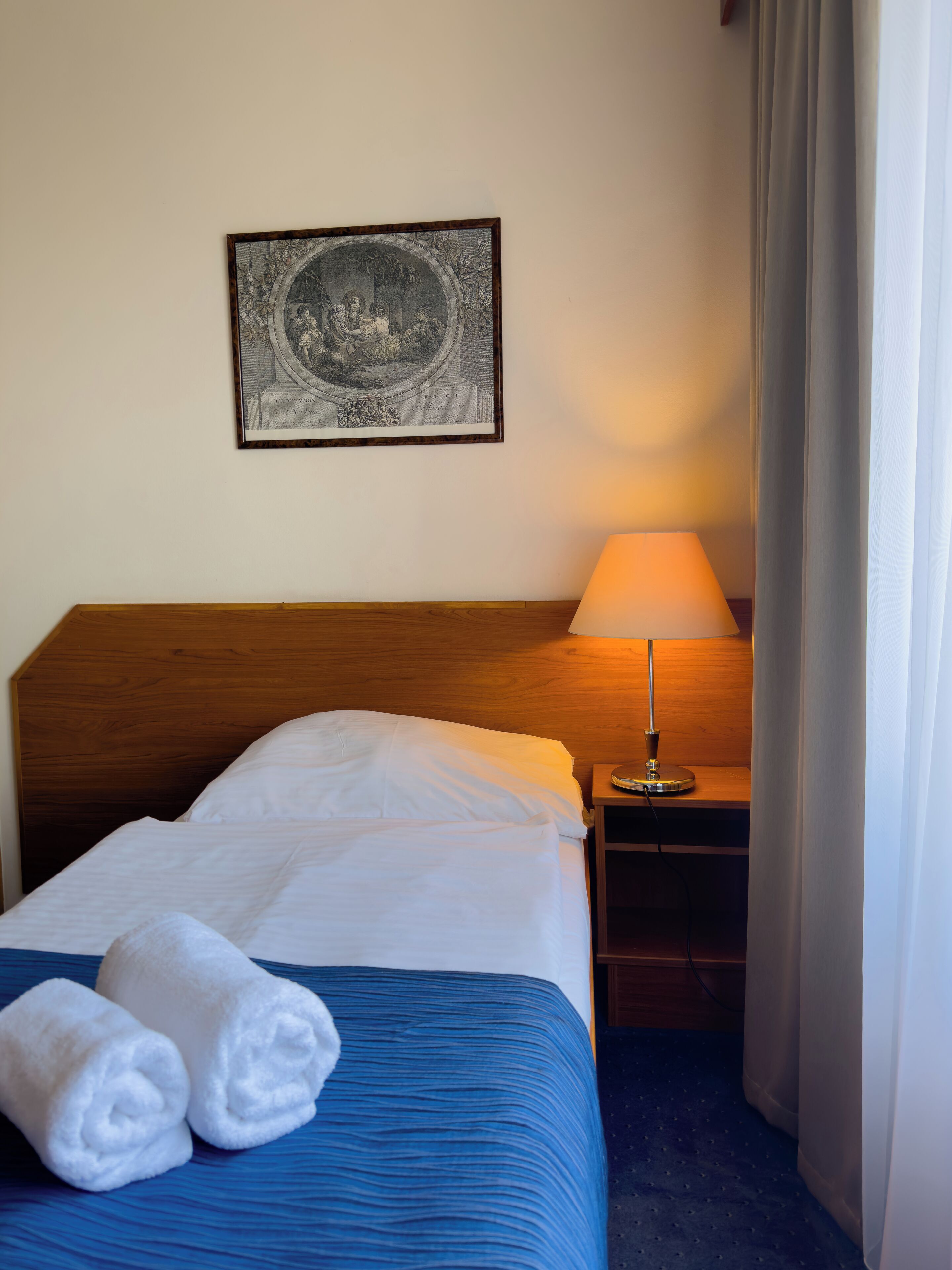 A neatly made bed with white linens and blue bedding, a bedside lamp, and a framed artwork on the wall.