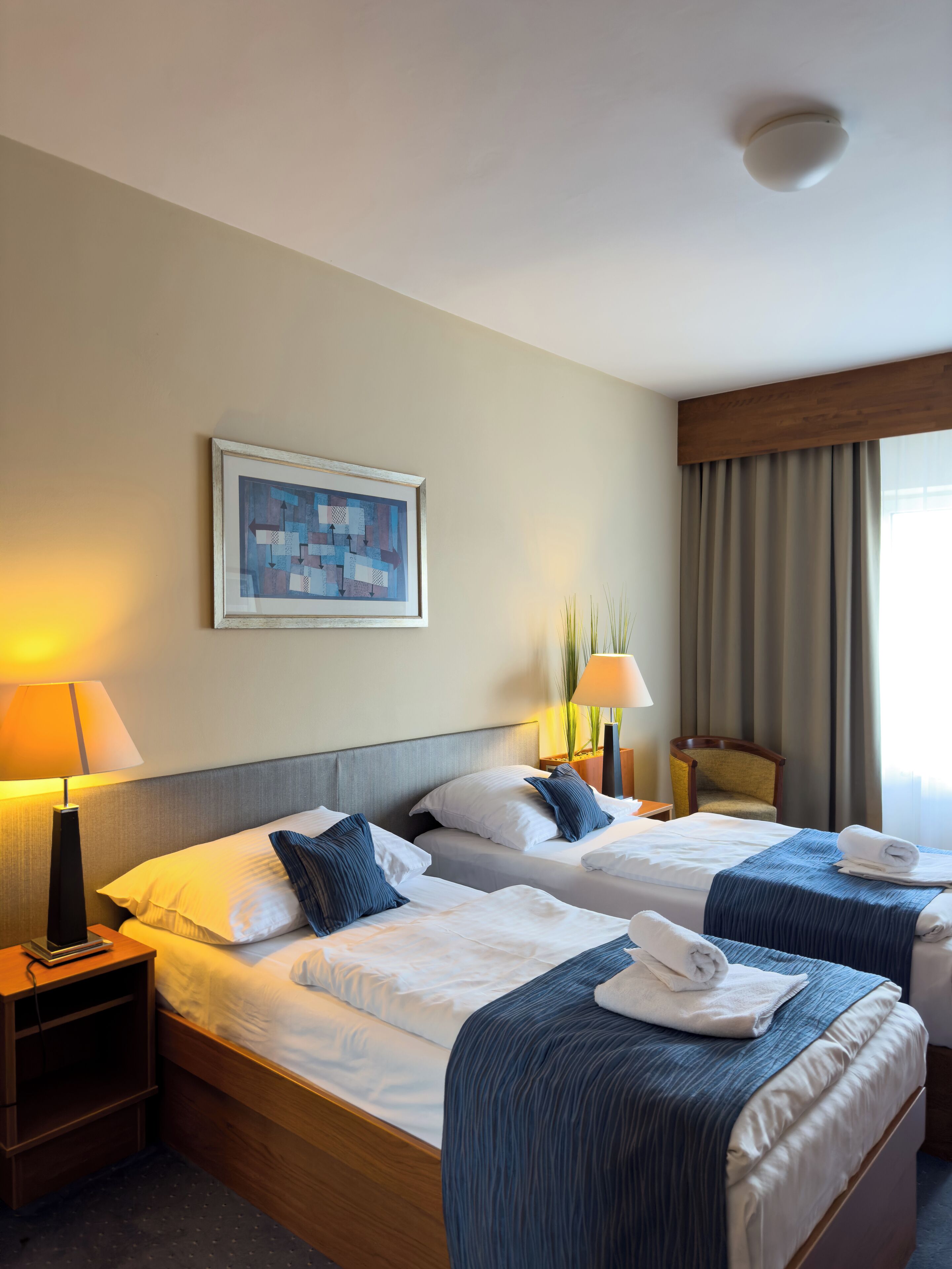 A hotel room with two beds, a framed artwork on the wall, a lamp, and a chair.