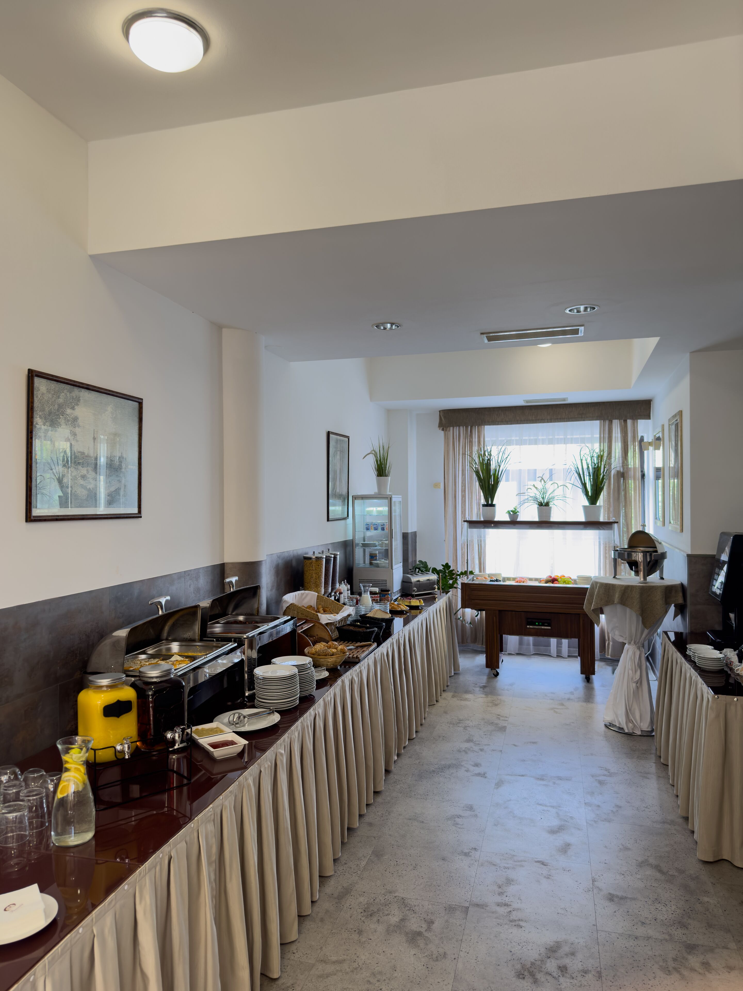 Montaje de bufé con varias estaciones de comida, una mesa larga cubierta con mantel y una ventana con cortinas al fondo.