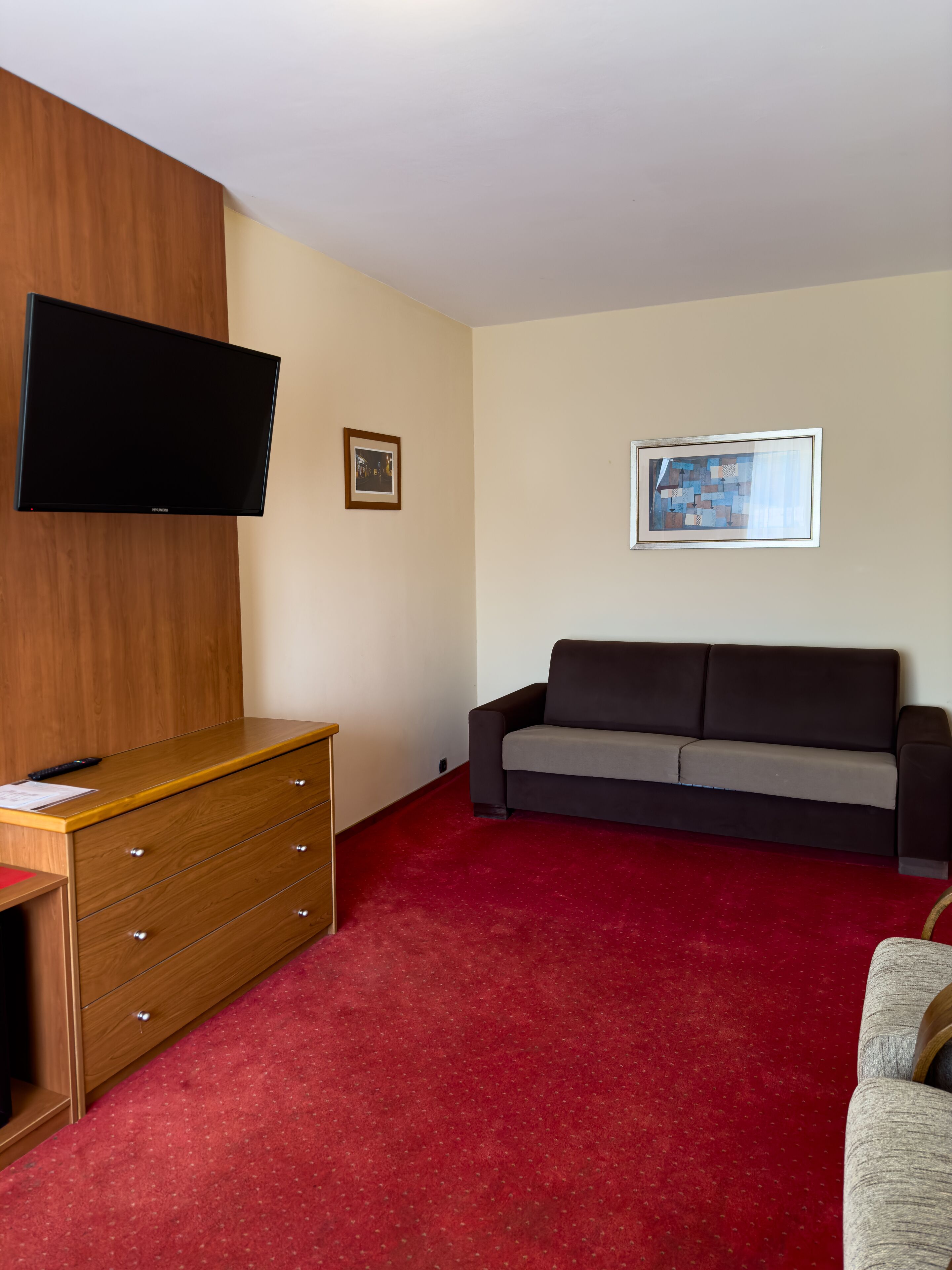 Une pièce comprenant une commode en bois, un canapé gris, une moquette rouge et un téléviseur à écran plat fixé au mur.