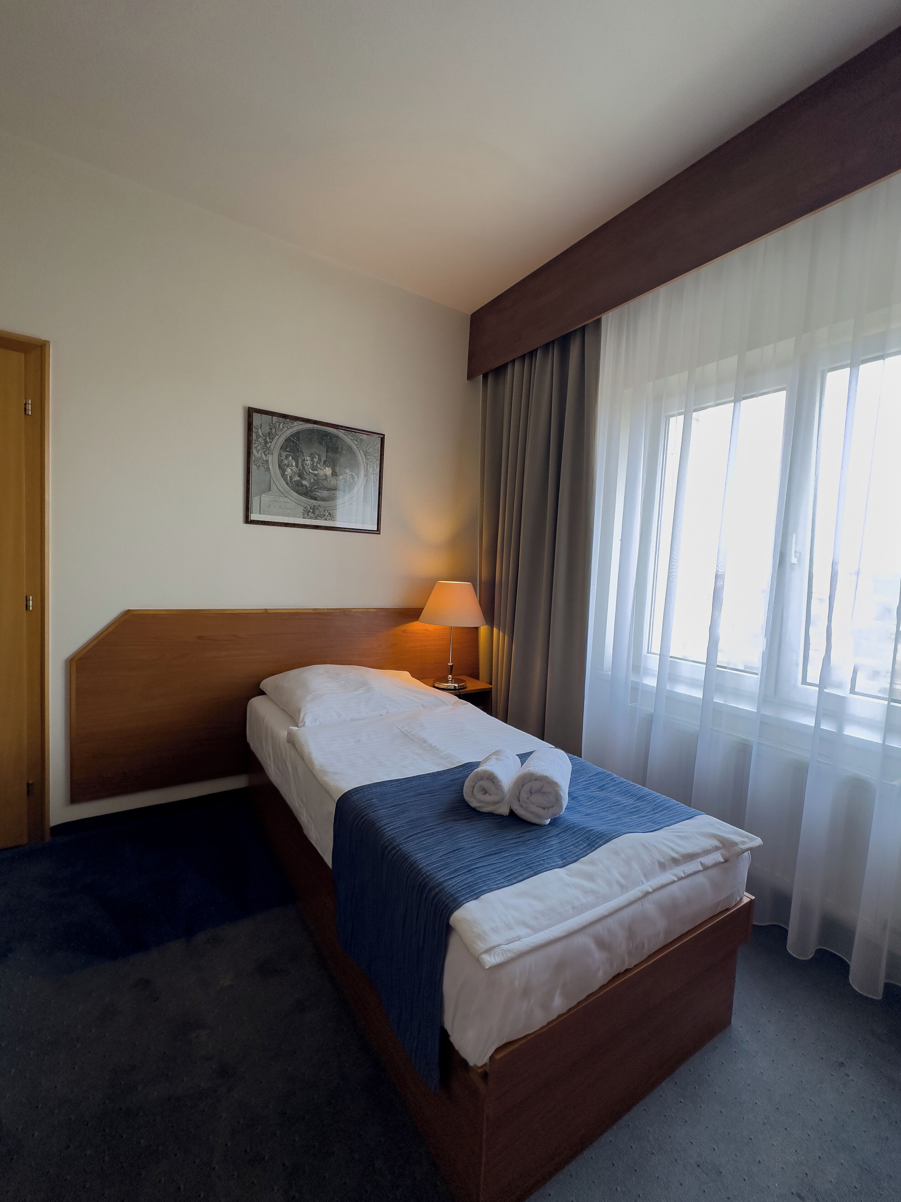 Habitación de hotel con cabecera de madera, cama con sábanas blancas y manta azul, lámpara de noche, un cuadro en la pared y ventana con cortinas traslúcidas.