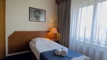Habitación de hotel con cabecera de madera, cama con sábanas blancas y manta azul, lámpara de noche, un cuadro en la pared y ventana con cortinas traslúcidas.
