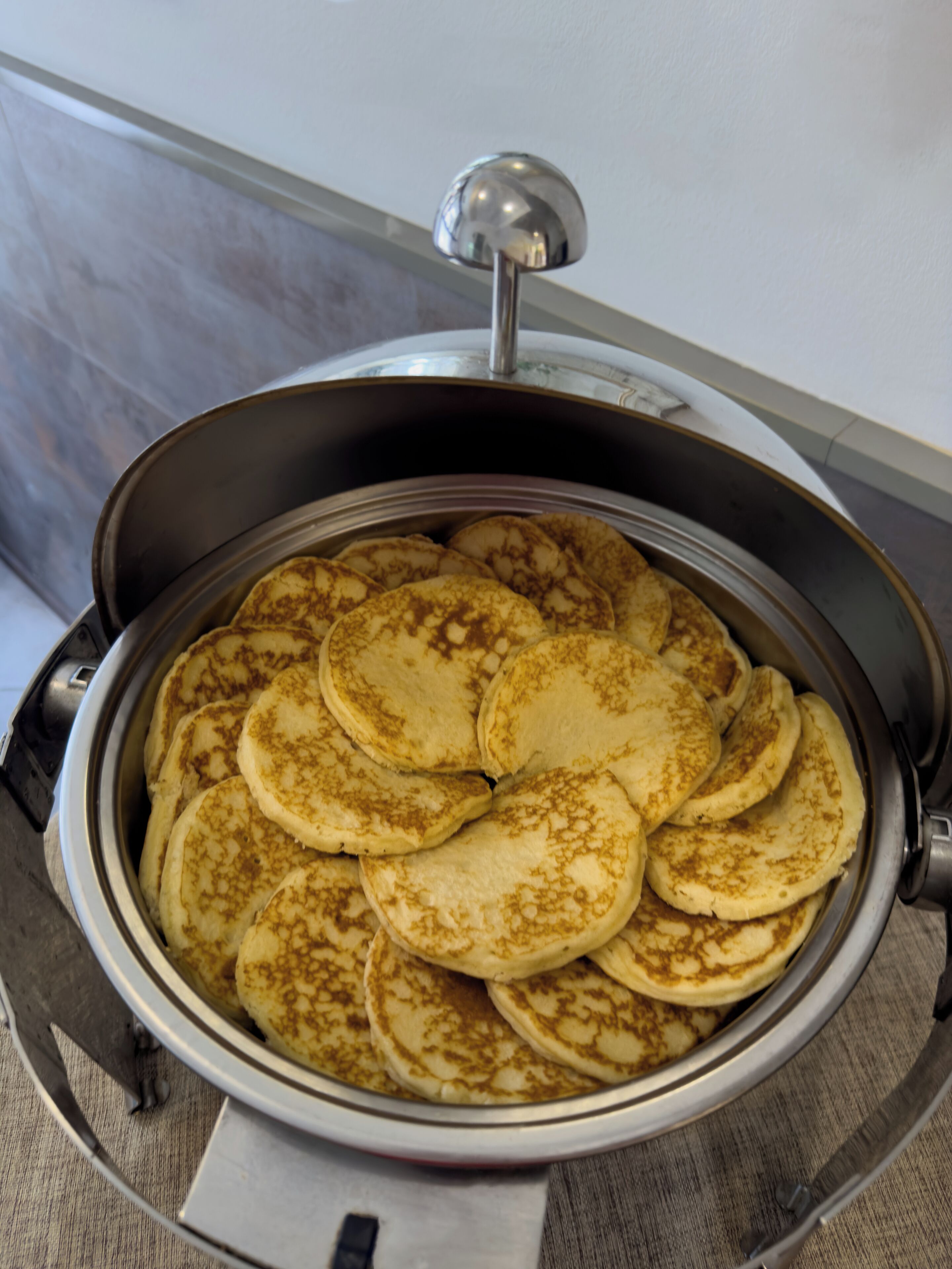 A round, stainless steel appliance with a dome-shaped lid, containing several pancakes stacked inside.