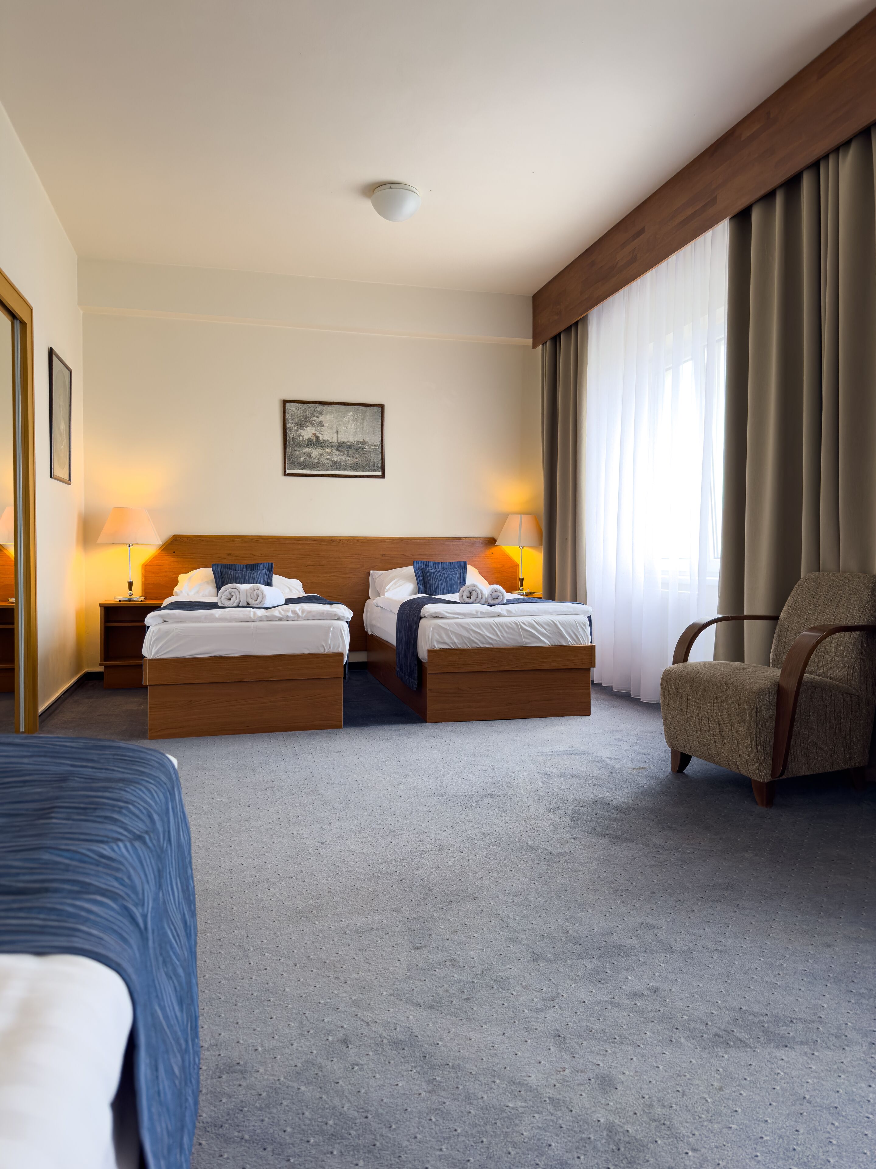 A hotel room with two beds, a chair, a painting on the wall, and a window with curtains.