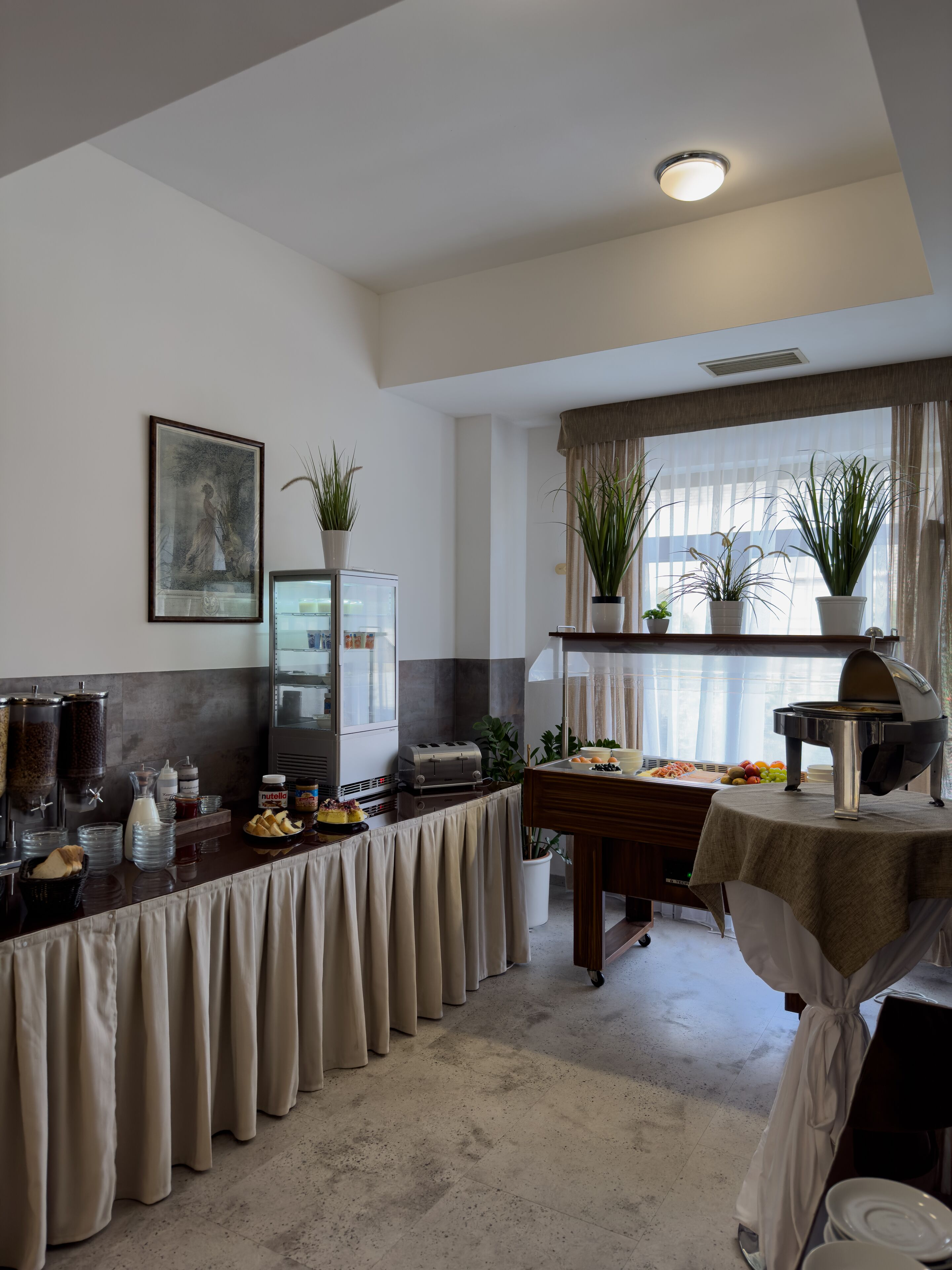 Una barra de desayuno con una cafetera, un tostador y una variedad de alimentos. Hay plantas en macetas y un cuadro en la pared.