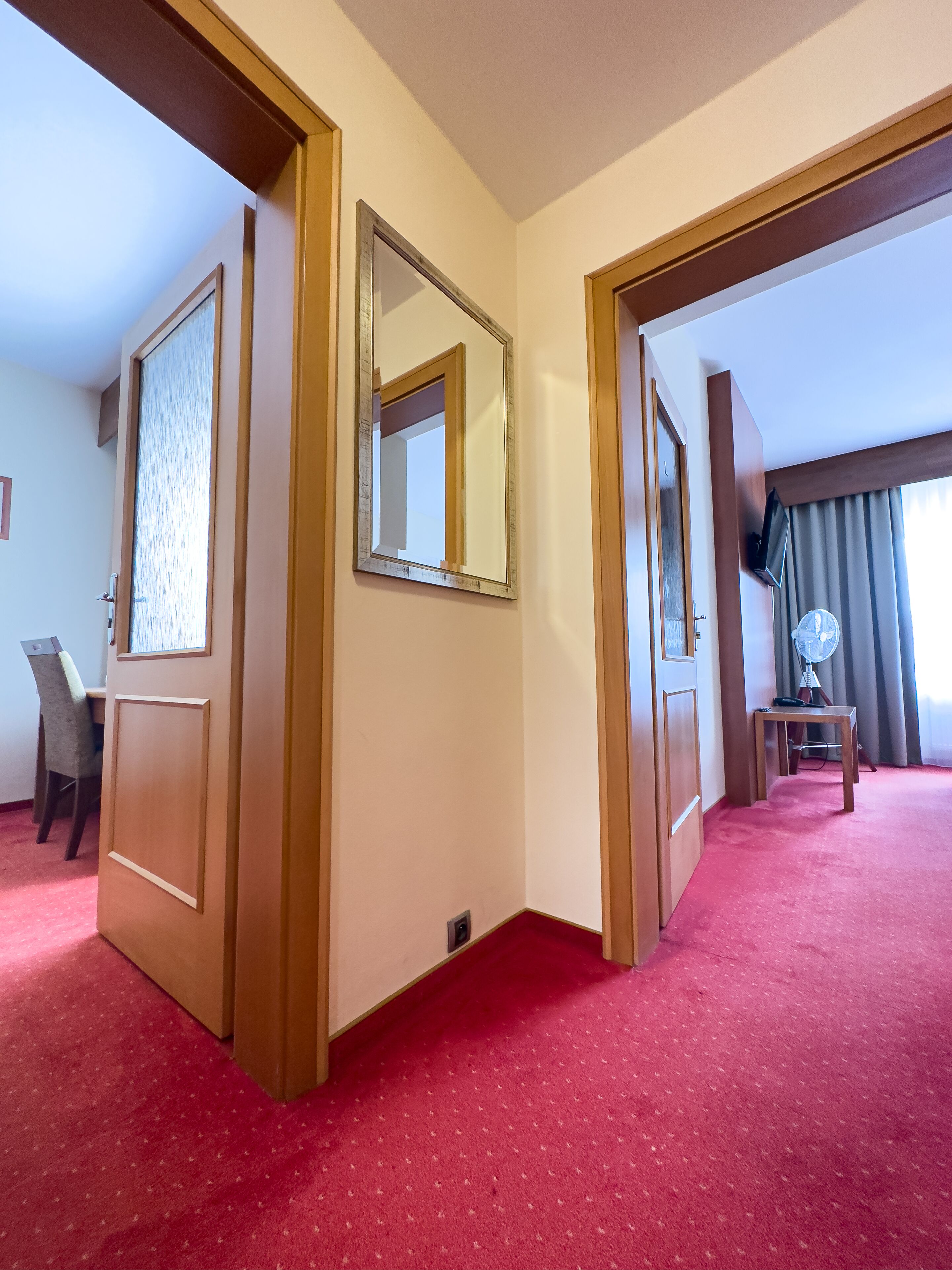 A hotel room with a red carpet, a wooden door, a mirror, a desk, and a chair.
