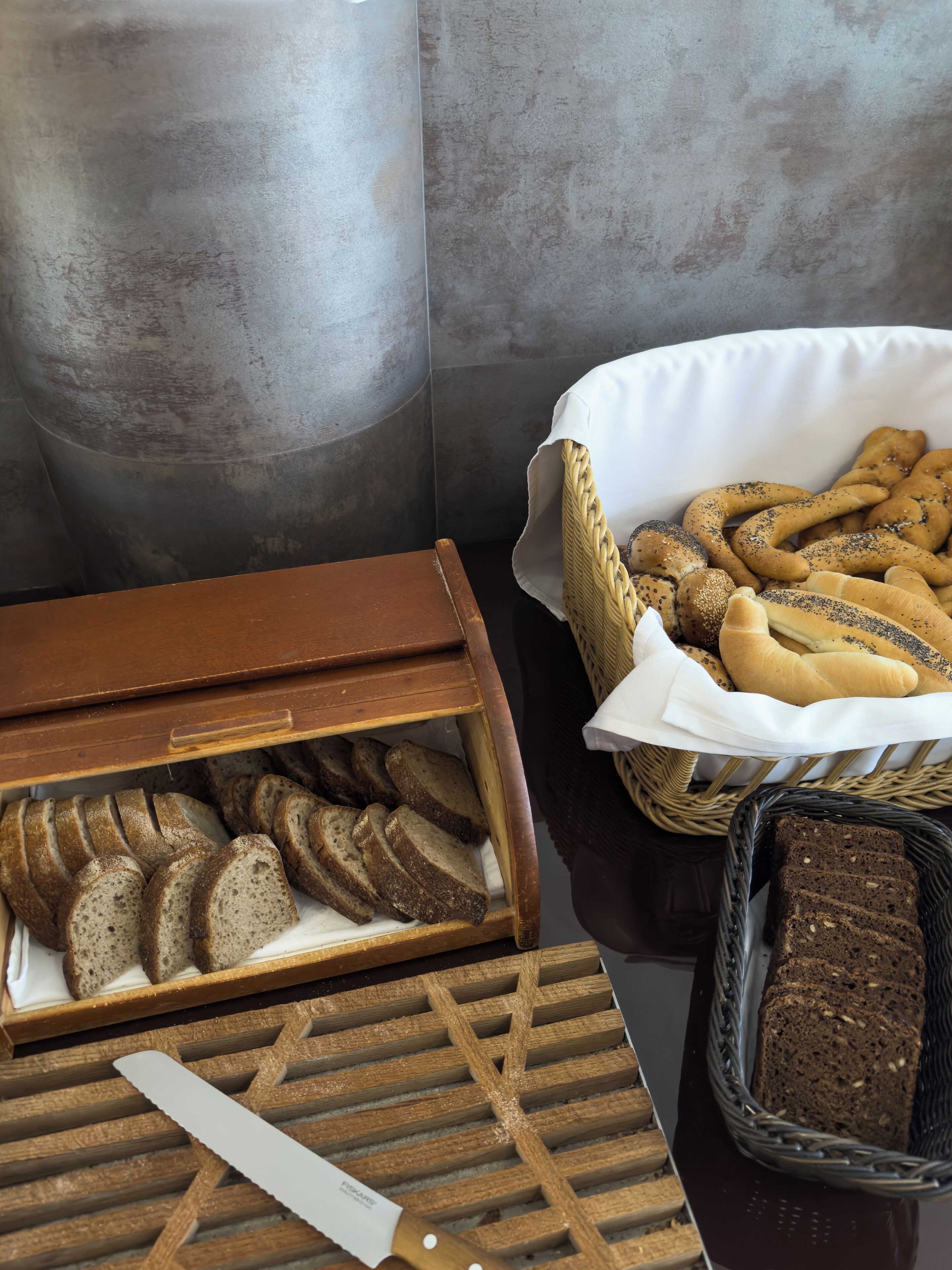 Una caja de madera para pan con pan en rebanadas, una cesta de mimbre con diferentes panes y una barra de pan oscuro sobre una mesa.