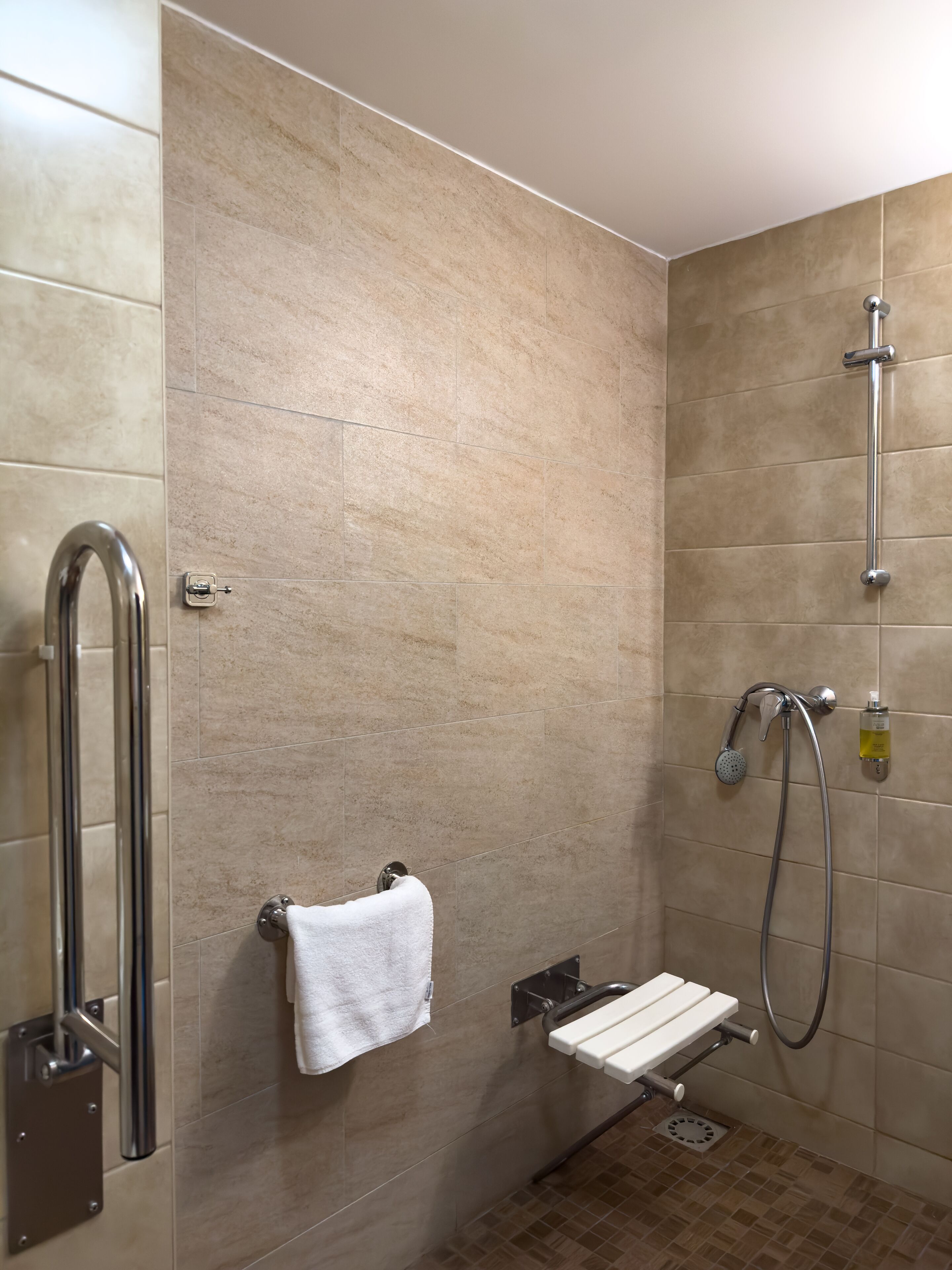 Un baño moderno con azulejos color beige, cabina de ducha de vidrio, una toalla blanca, cabezal de ducha de mano y dispensador de jabón.
