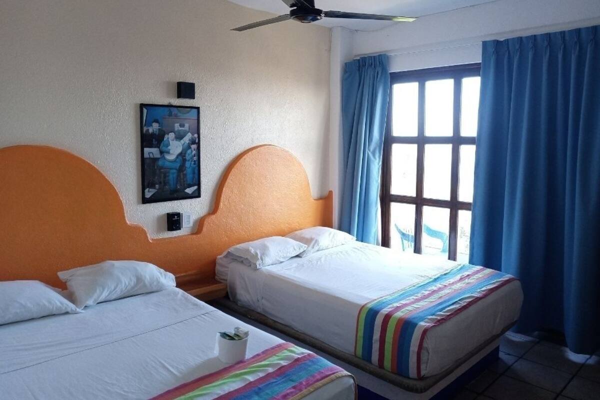 Habitación de hotel con dos camas, ventilador de techo, ventana con cortinas azules y un cartel en la pared.
