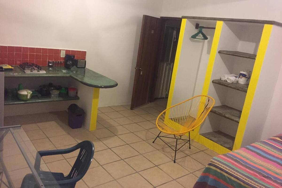 Una habitación compacta con kitchenette, una silla, una cama y un armario con detalles amarillos.