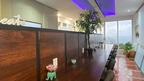 A reception area with a wooden reception desk, a large potted plant, and a view of the sea through large windows. - Pargo's Hotel (Puerto Escondido)