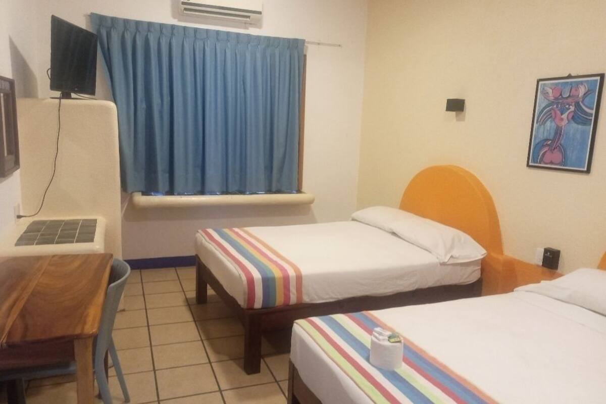 Habitación de hotel con dos camas individuales, un escritorio de madera, una silla, una ventana con cortinas azules y un aire acondicionado de pared.
