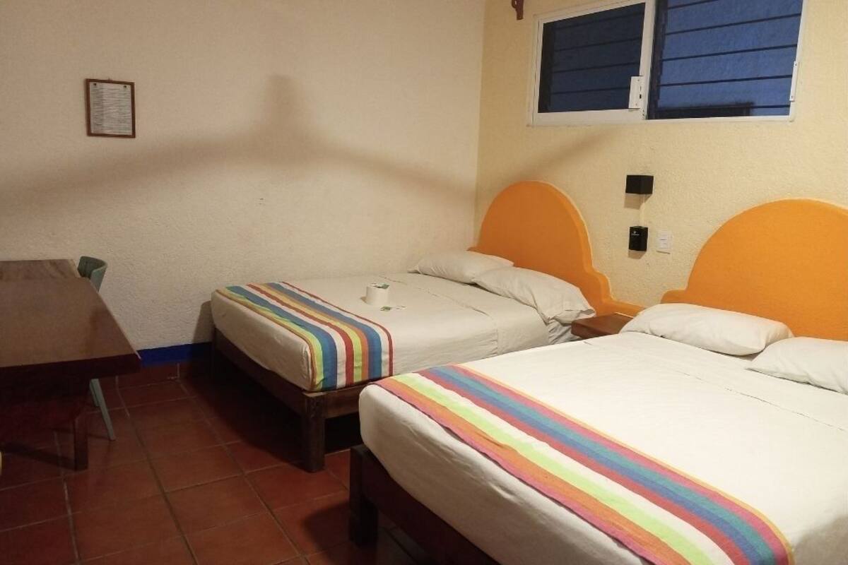 Una habitación con dos camas, cada una con un colorido edredón a rayas, una mesa de madera y una ventana con contraventanas azules.