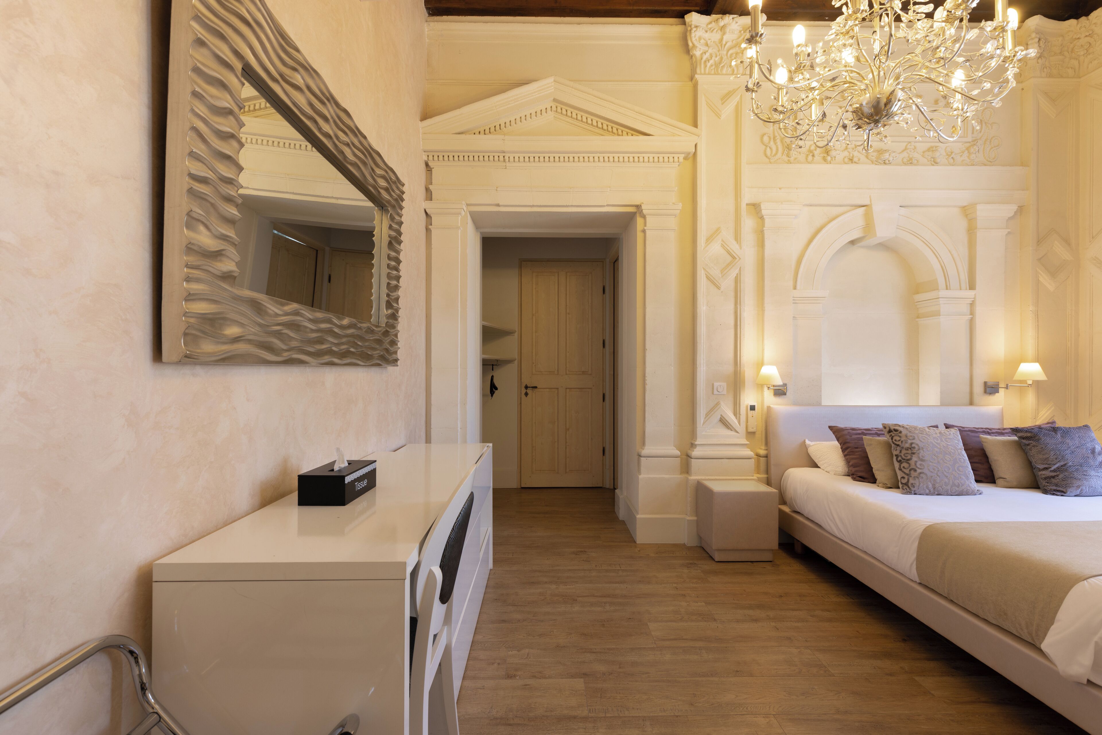 Une chambre d’hôtel comprenant un lit, une commode, un miroir, un lustre et une porte donnant sur une autre pièce.