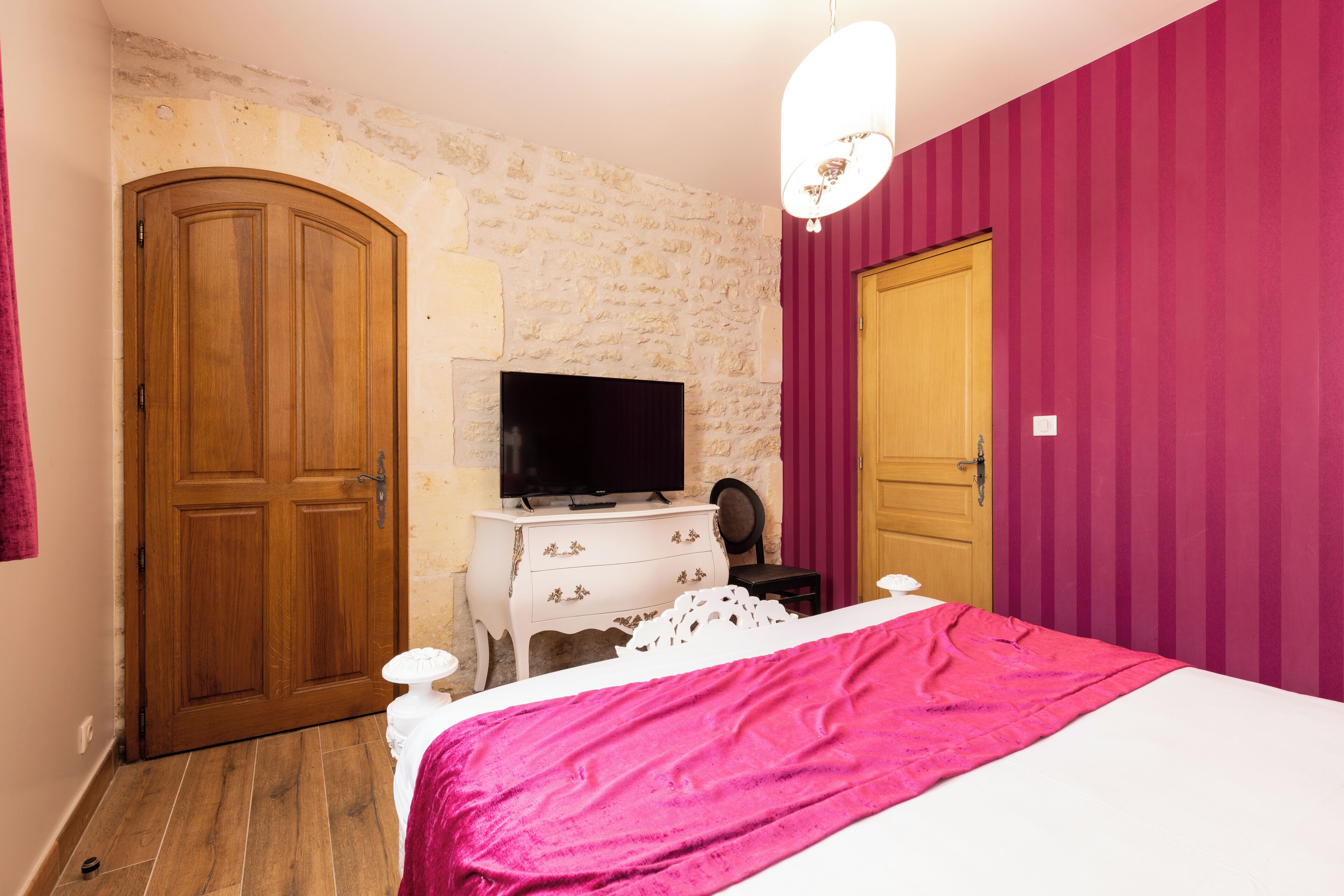 Un dormitorio con pared de piedra, armario de madera, televisor sobre una cómoda y cama con manta rosa.