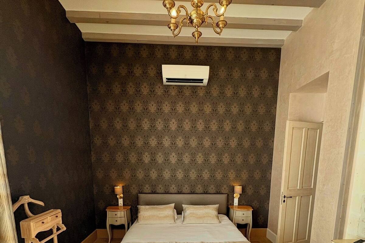 A bedroom with a large bed, wooden floor, dark patterned wallpaper, a chandelier, bedside tables, and a wardrobe.