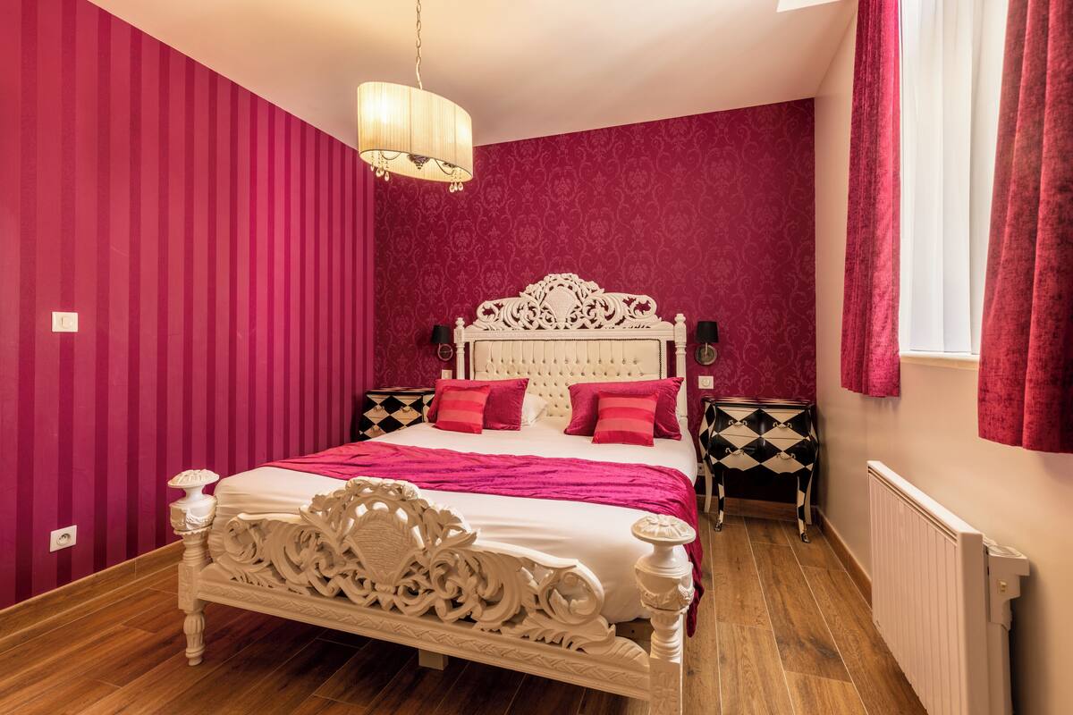 A bedroom with a large bed, a chandelier, a dresser, and a radiator.