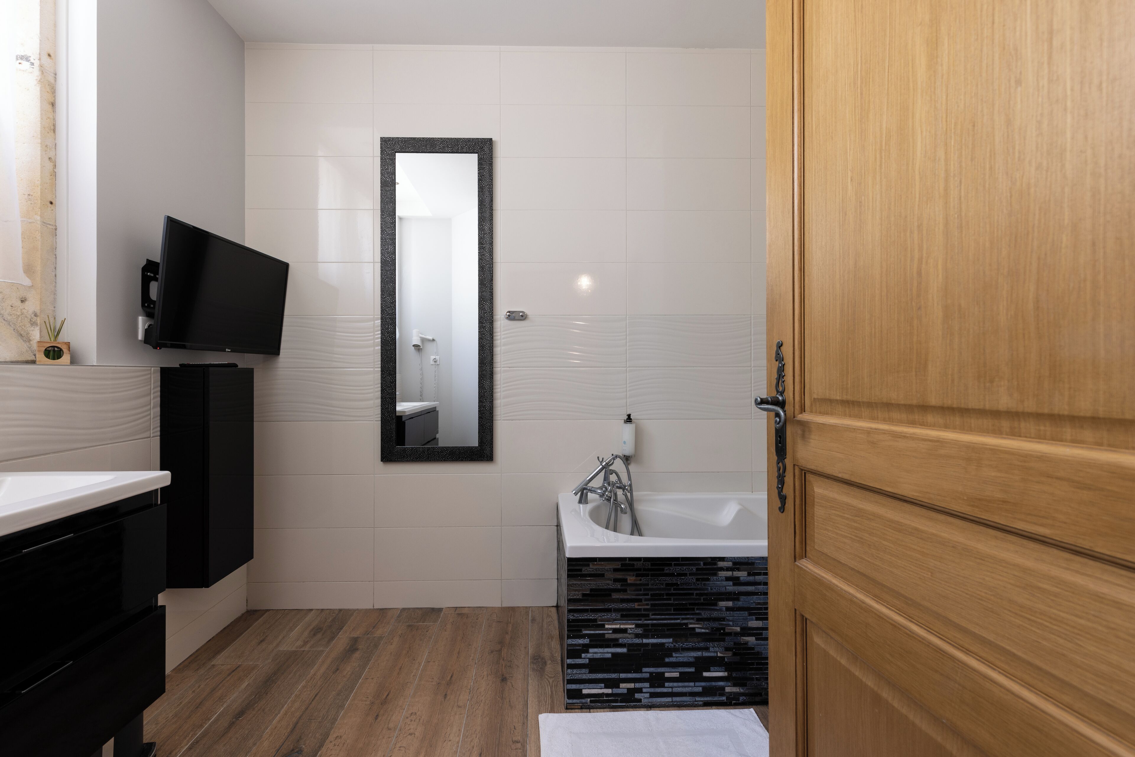 A modern bathroom with a wooden door, a large mirror, a black cabinet, a white sink, and a black and white tiled floor.
