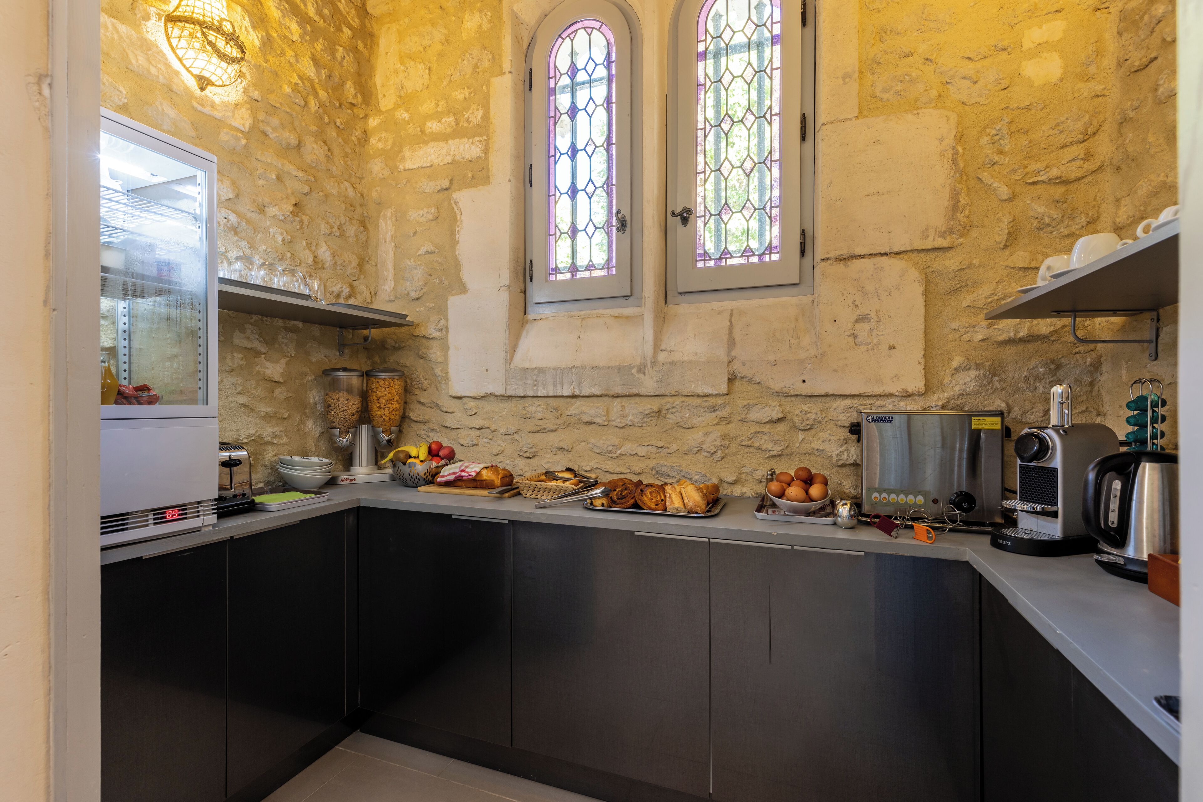 A kitchen with stone walls, two windows, a coffee machine, and various food items on the counter.
