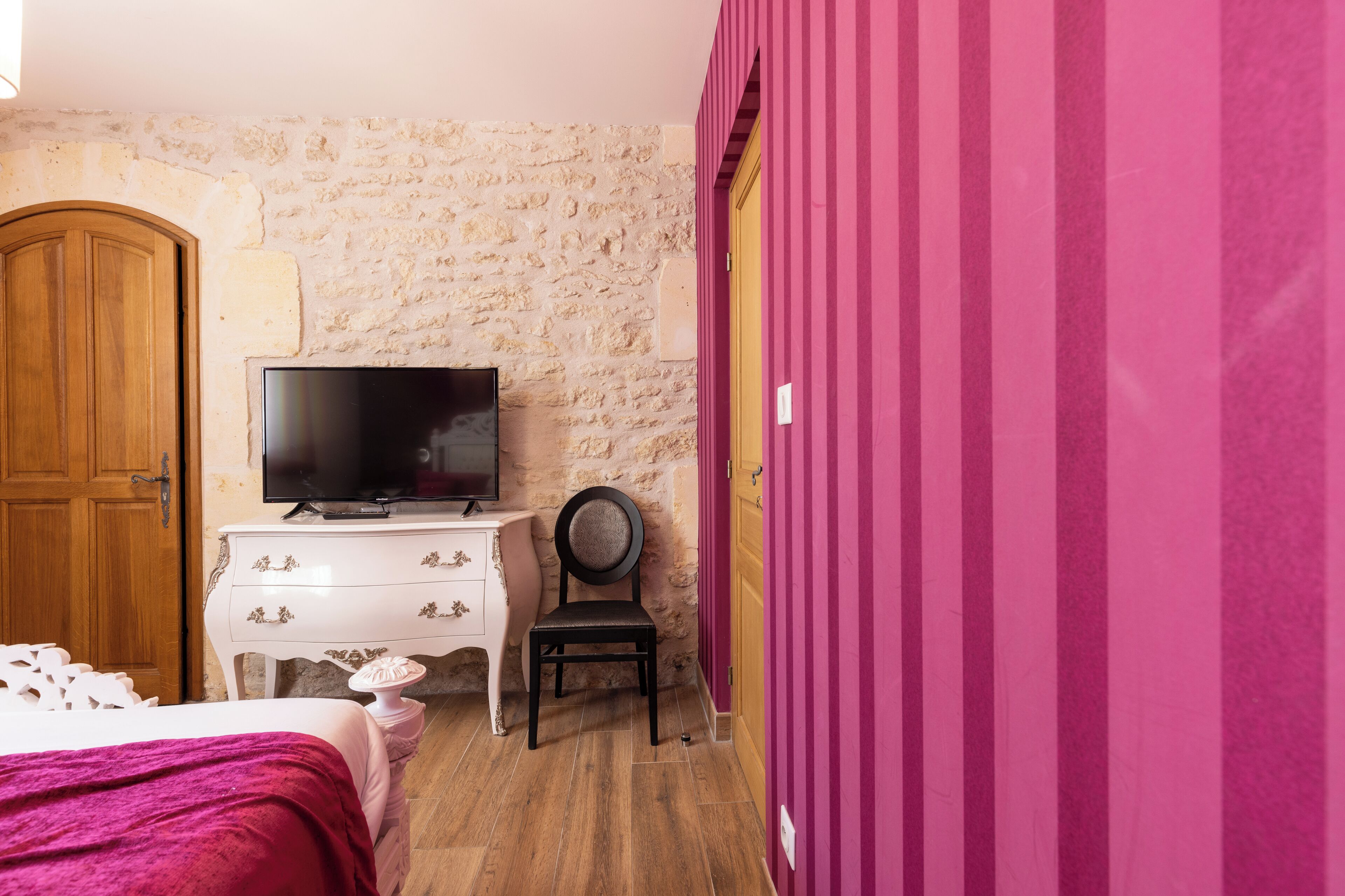 A bedroom with a stone wall, a wooden door, a TV on a white dresser, a black chair, and a pink wall.