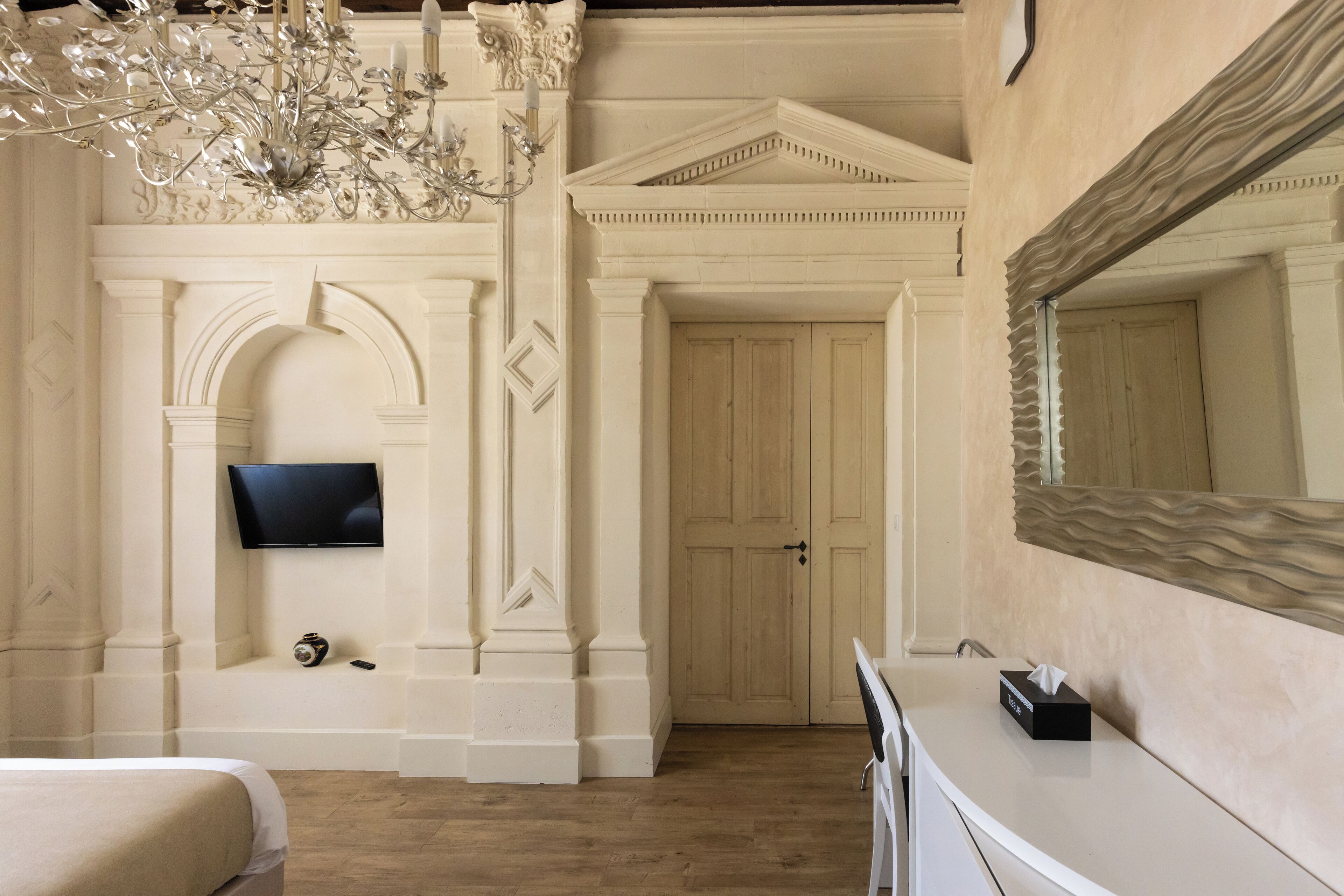 A hotel room with a large bed, a flat-screen TV mounted on the wall, a chandelier, and a large mirror.