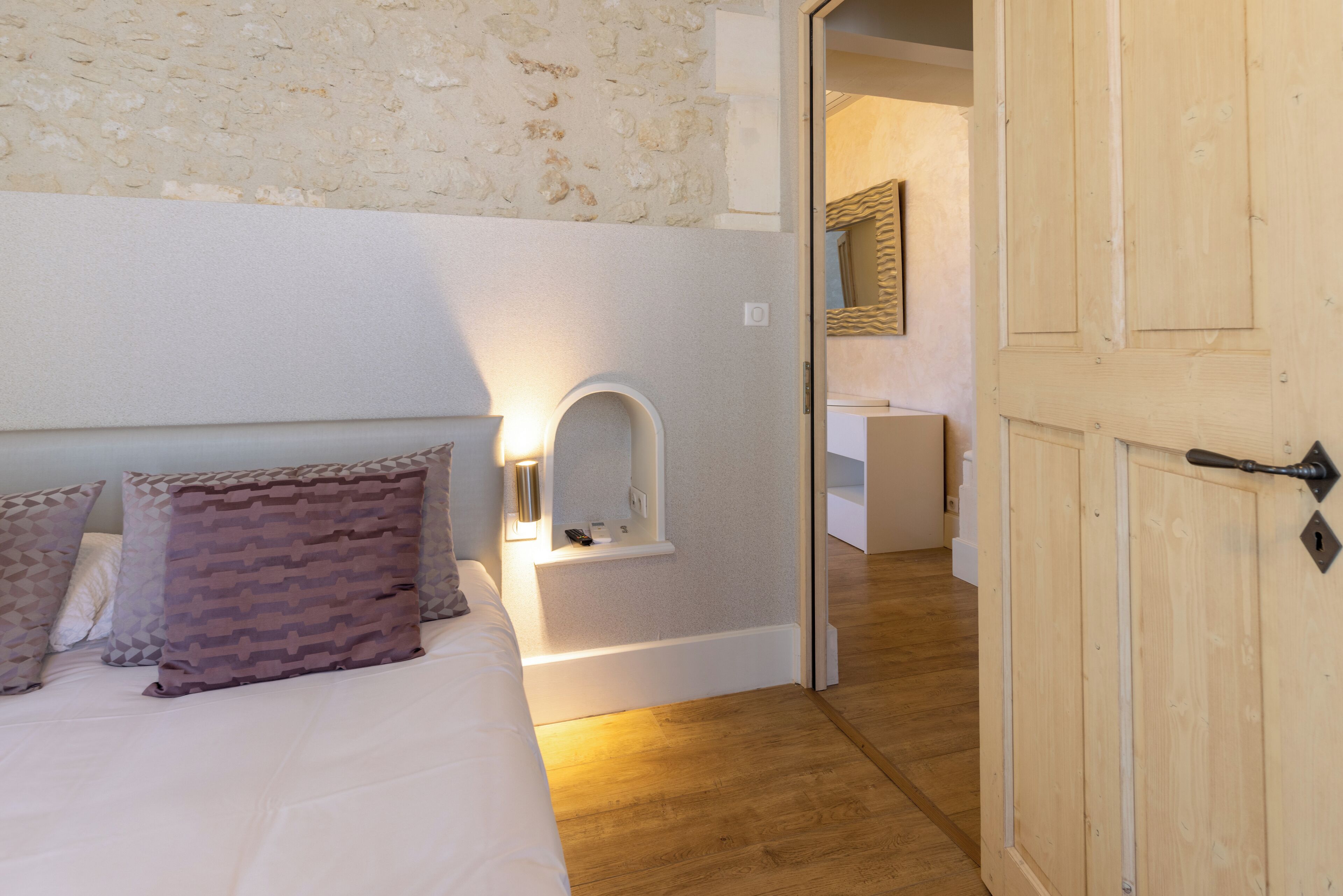 Une chambre à coucher avec un lit, des oreillers, une table de chevet avec une lampe, une porte en bois et un mur en pierre.