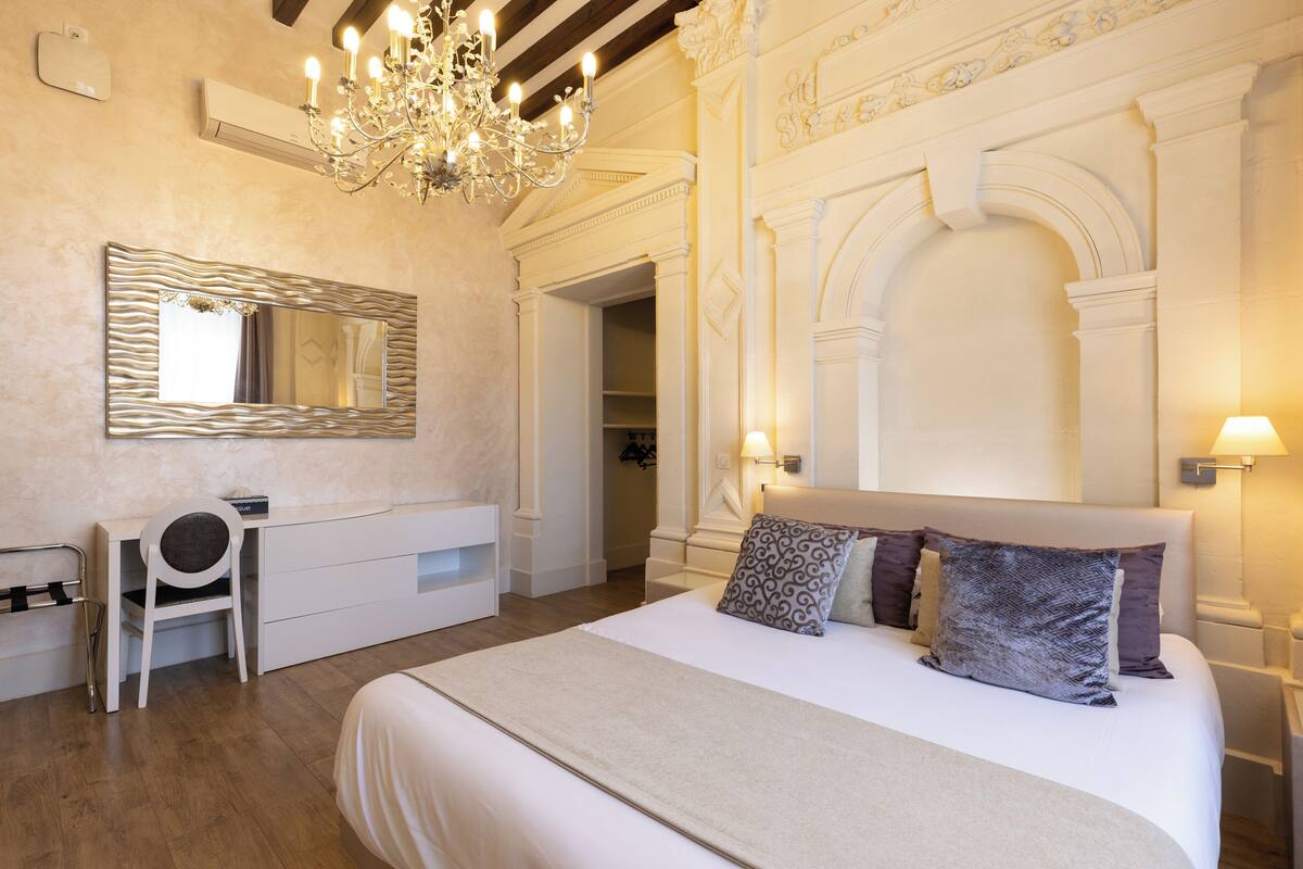 A bedroom with a large bed, a desk, a chair, a chandelier, and a mirror.