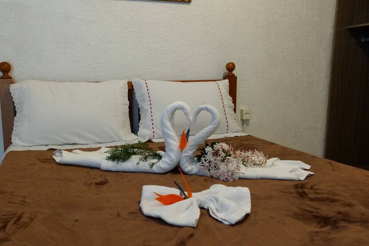 Een bed met een bruine deken, twee witte kussens en decoratieve handdoeken in de vorm van zwanen met bloemen.
