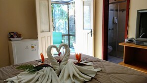 Une chambre d’hôtel avec un lit agrémenté d’une décoration en serviettes en forme de deux cygnes, un réfrigérateur blanc, un téléviseur posé sur un meuble en bois, et une salle de bain visible à travers une porte ouverte.